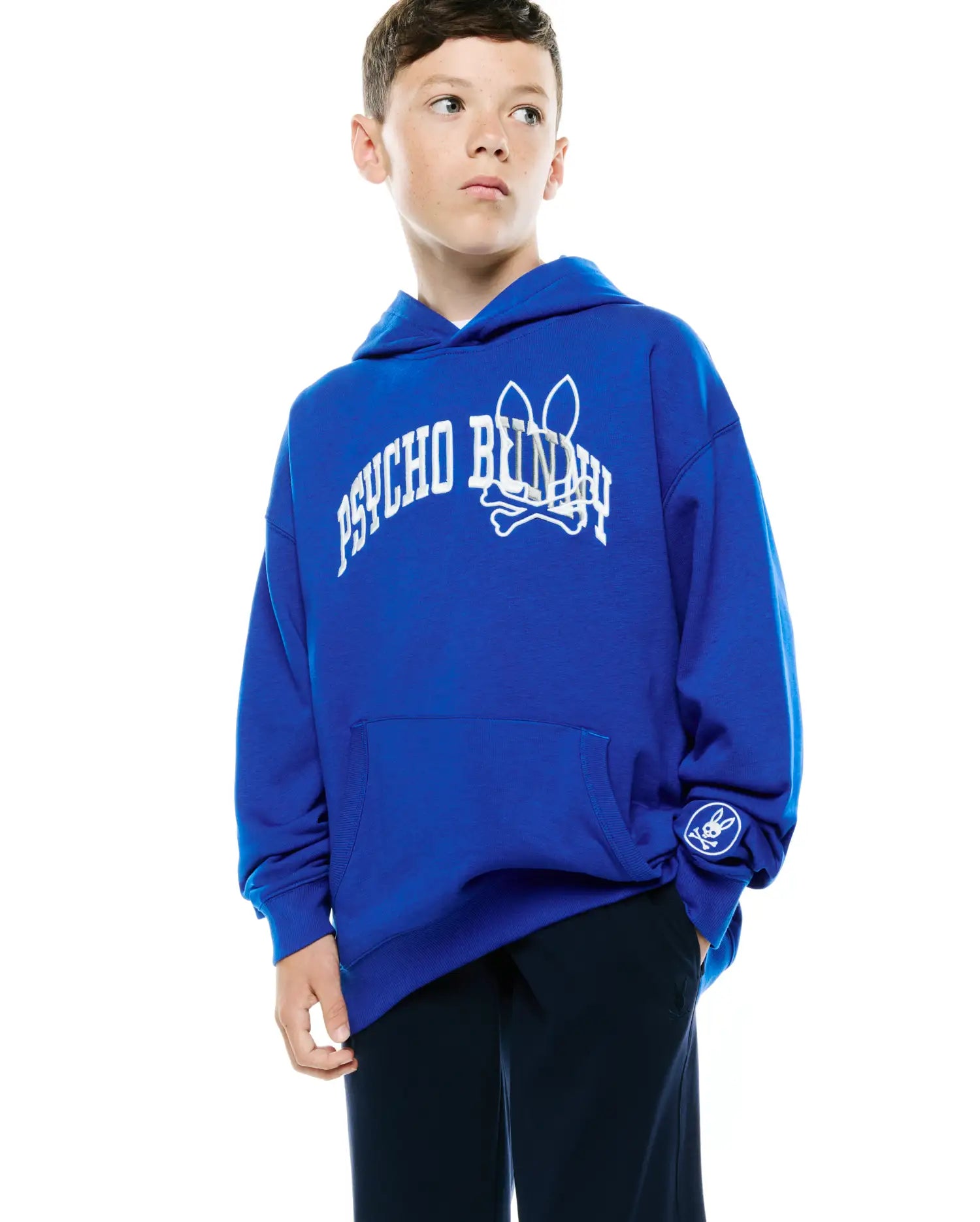 Kids Nathaniel Relaxed Hoodie in royal blue, featuring a large white arched Psycho Bunny text and skull logo on the chest, a small white circular skull logo on the left sleeve, and a kangaroo pocket. Smooth jersey fabric.