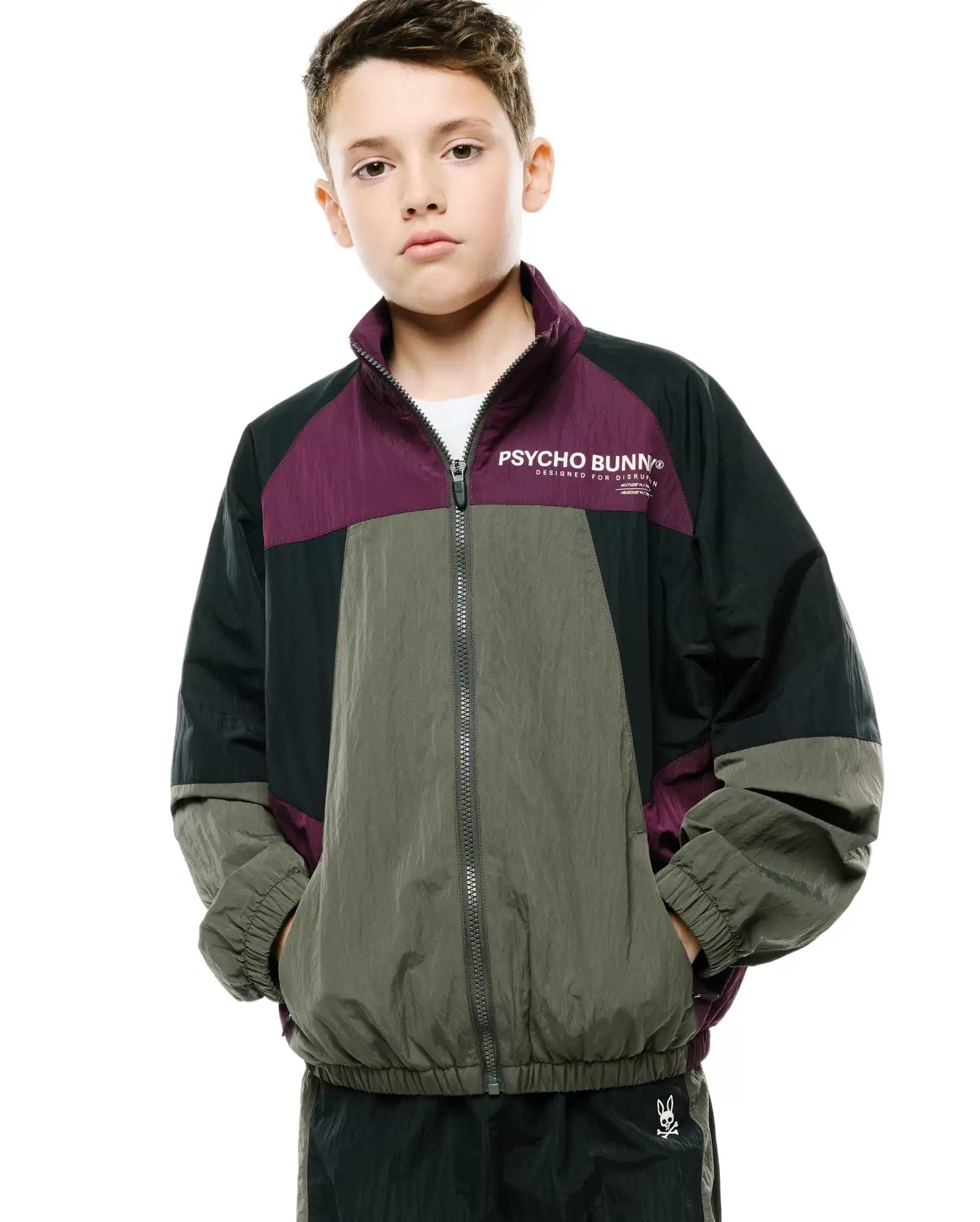 Kids Alder Nylon Colorblock Jacket, a relaxed fit full-zip jacket in crinkly nylon, featuring color blocks of deep purple, dark green, and olive green, with a white Psycho Bunny text logo and slogan on the chest.