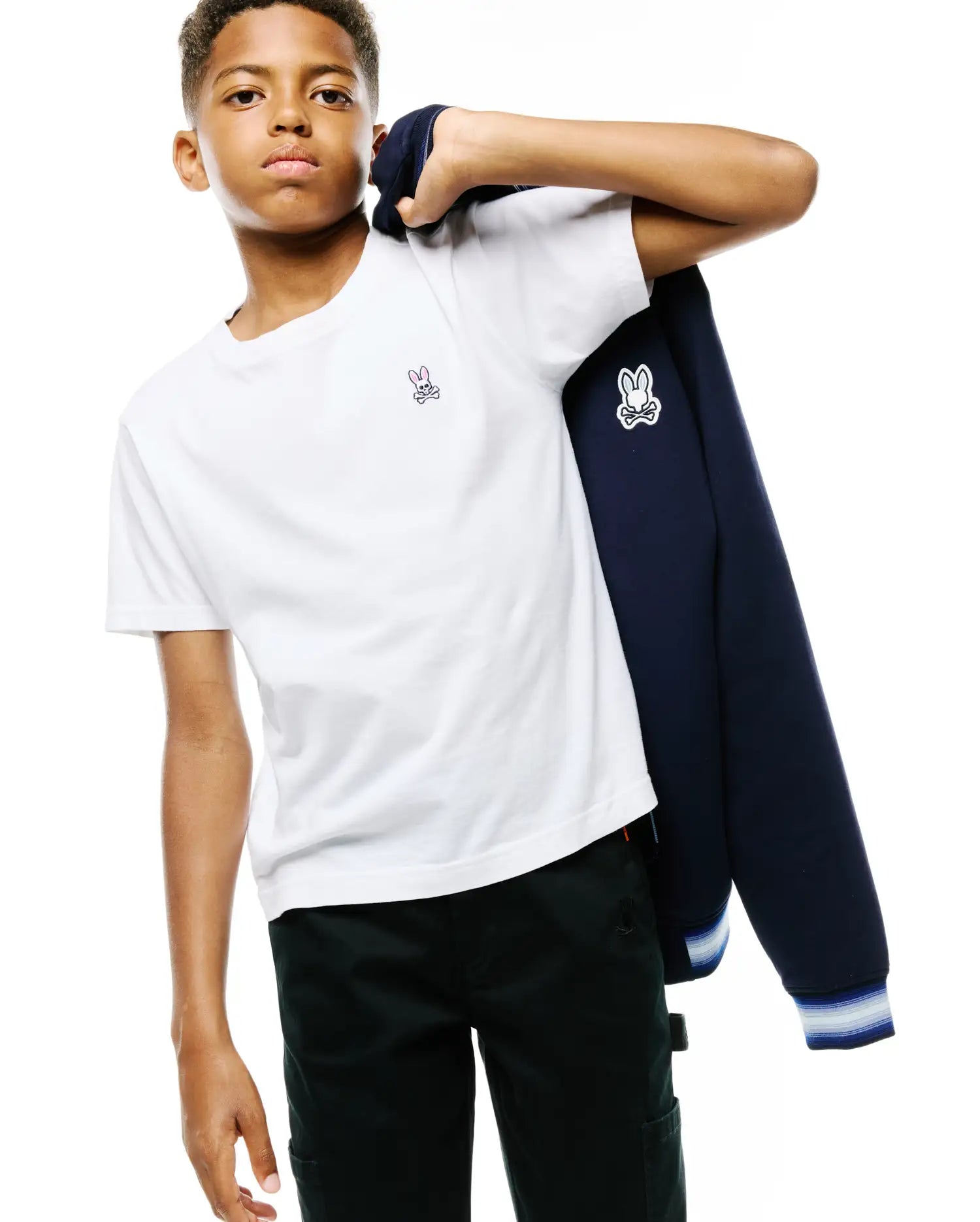 White jersey t-shirt with small embroidered pink and white Psycho Bunny logo, dark green pants, and a navy blue KIDS JAYDON TRACK JACKET with a large white and black Psycho Bunny logo on the sleeve and striped cuffs.