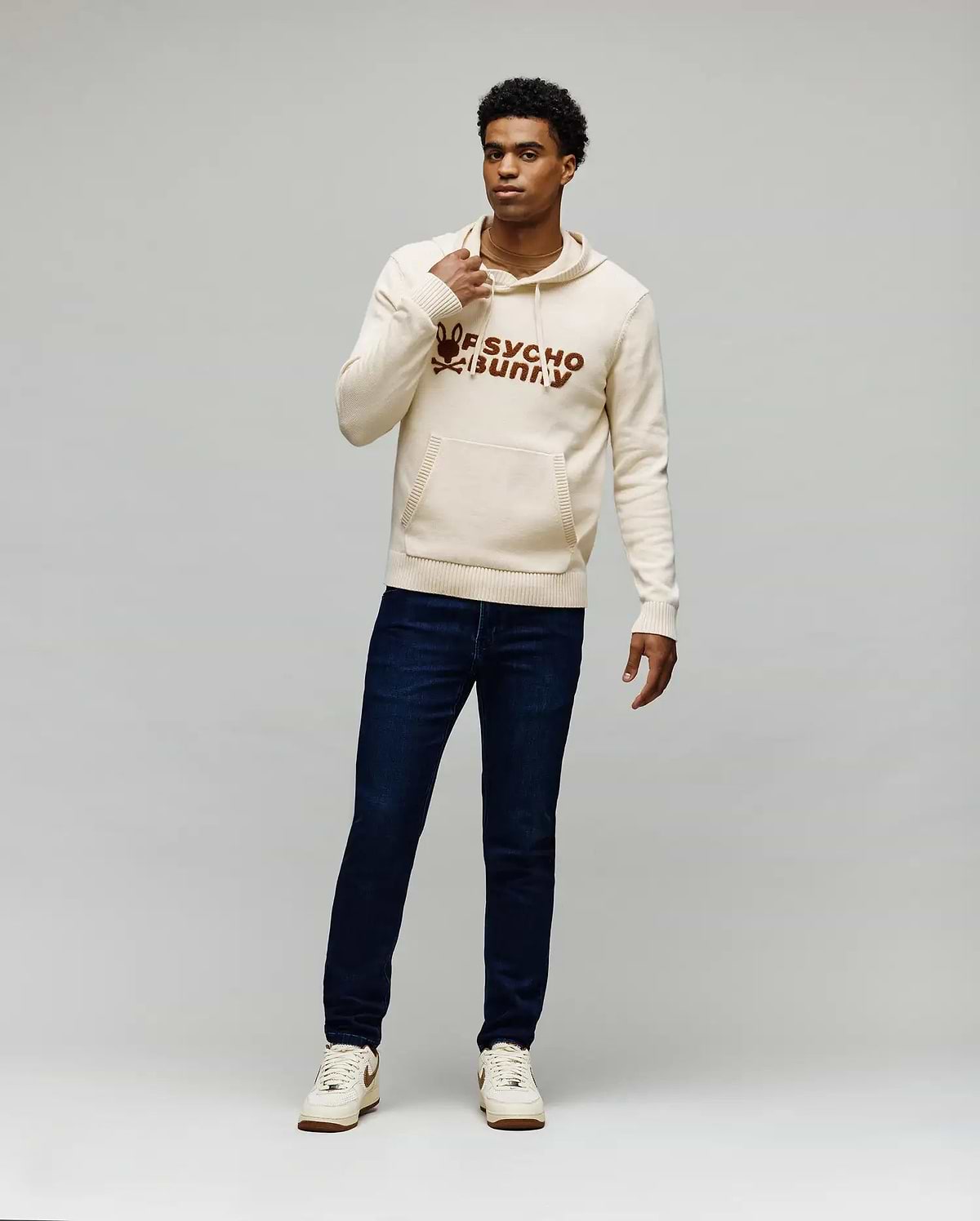 Cream knitted hoodie with a large brown embroidered Psycho Bunny skull logo and text on the chest, kangaroo pocket, and ribbed details, paired with dark blue slim-fit Mens Emerson Denim Pant.