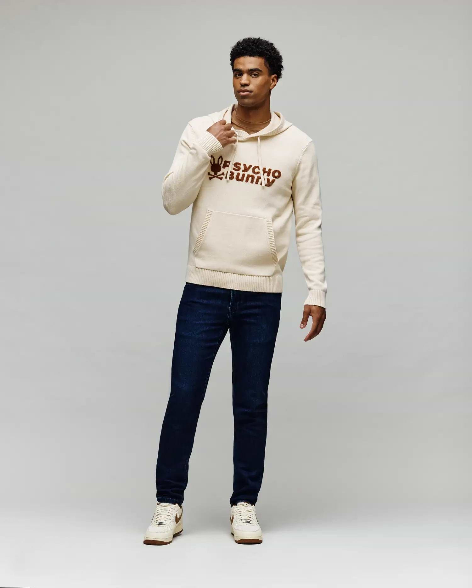 Cream knitted hoodie with a large brown embroidered Psycho Bunny skull logo and text on the chest, kangaroo pocket, and ribbed details, paired with dark blue slim-fit Mens Emerson Denim Pant.