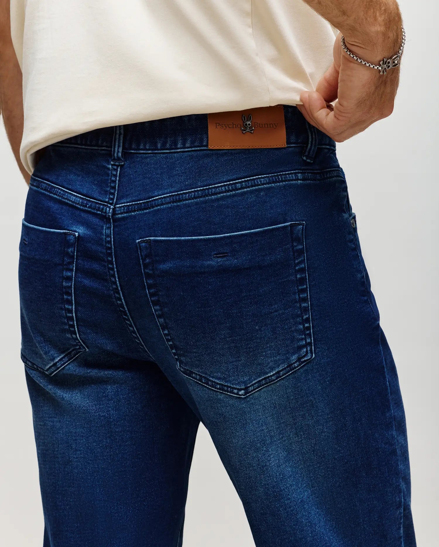 Mens Ace Knit Denim Pant in dark blue, classic fit, with subtle fading, back pockets, and a brown leather Psycho Bunny skull logo patch on the waistband.