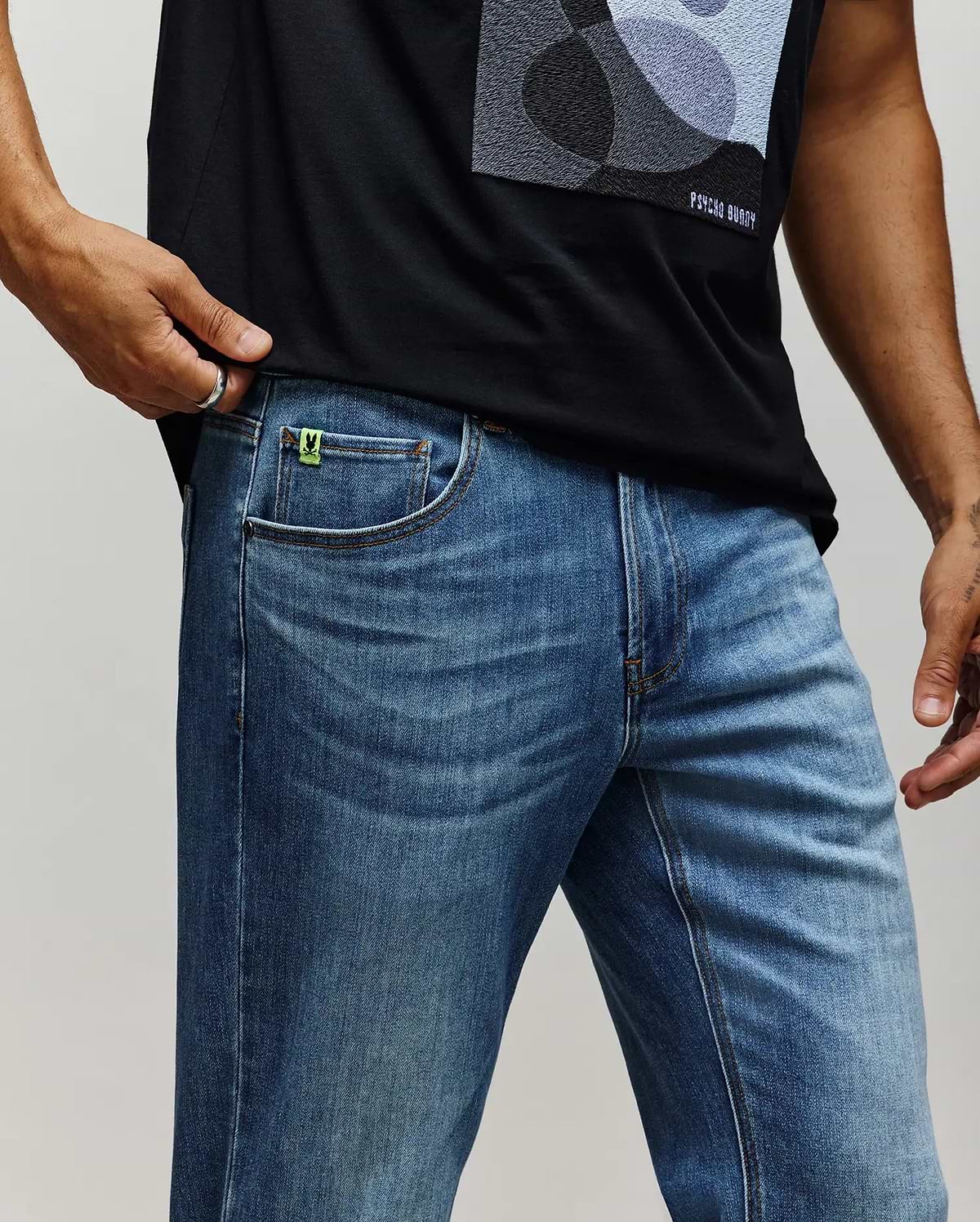 Black jersey t-shirt with abstract grey/blue chest graphic and white Psycho Bunny text, paired with medium wash Mens Emerson Denim Pant featuring a neon green bunny skull logo tag on the coin pocket.
