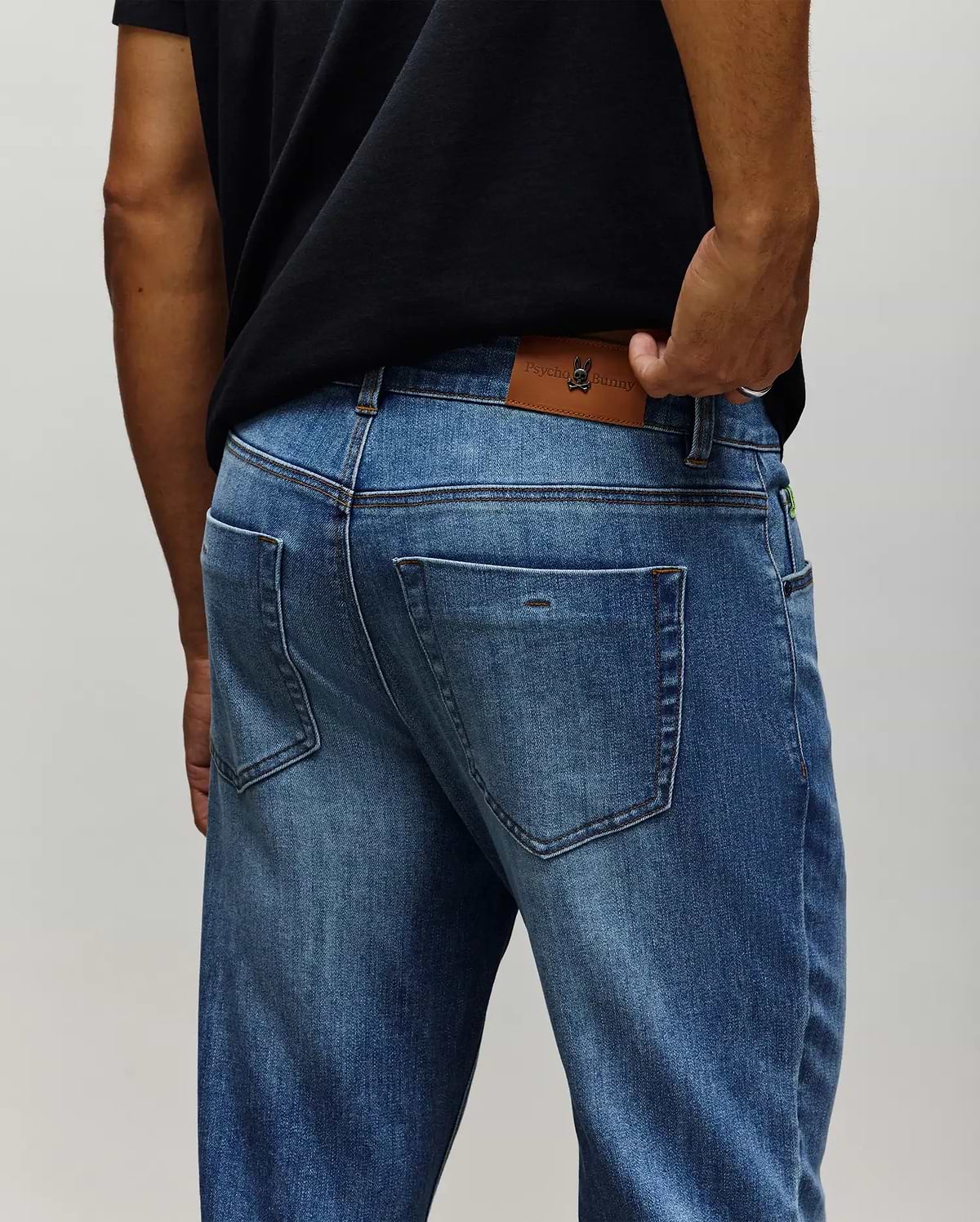 Mens Emerson Denim Pant in medium wash blue denim, featuring a classic fit, faded wash, contrast stitching, and a large brown leather Psycho Bunny skull logo patch on the back waistband with a small green tag.