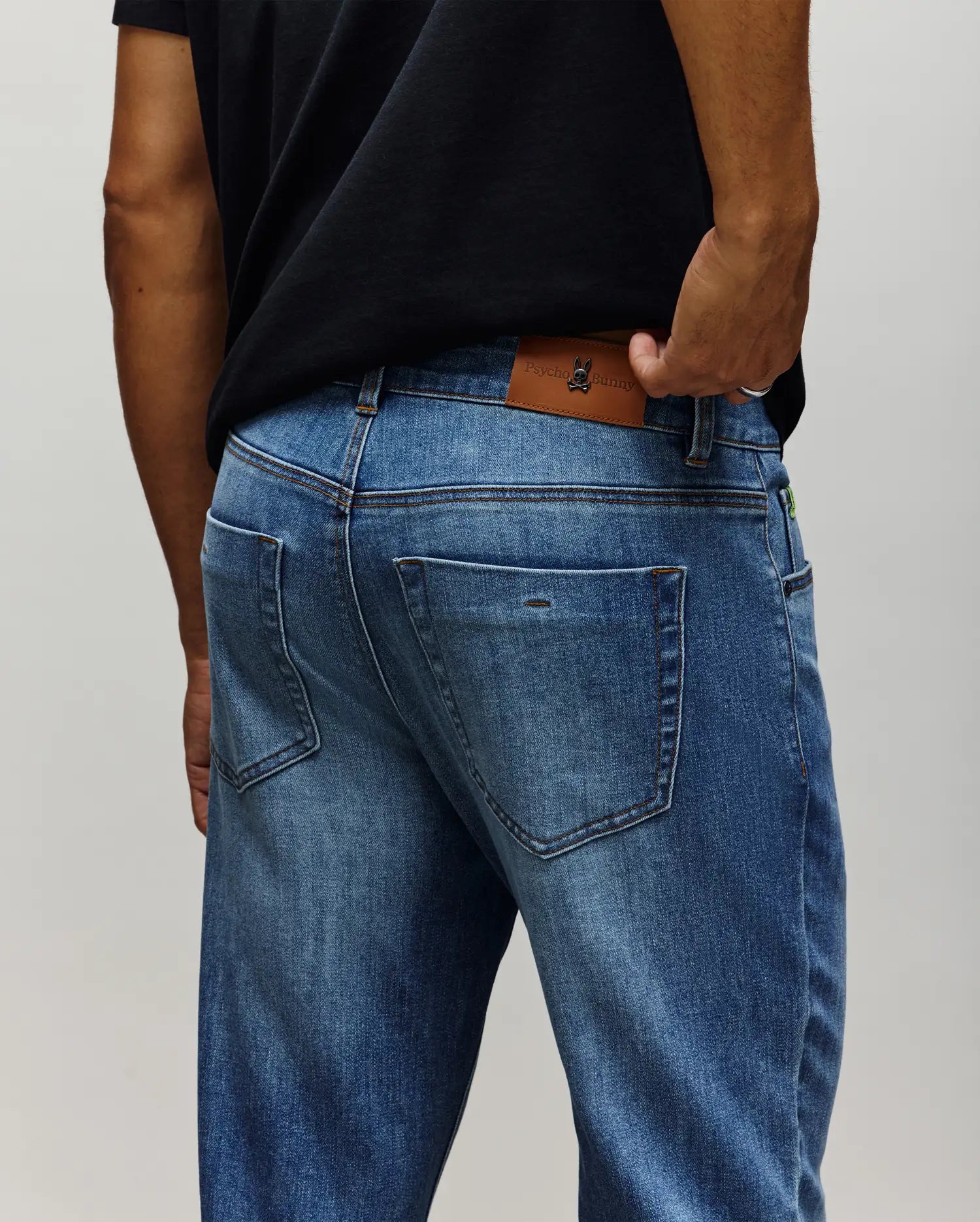 Mens Emerson Denim Pant in medium wash blue denim, featuring a classic fit, faded wash, contrast stitching, and a large brown leather Psycho Bunny skull logo patch on the back waistband with a small green tag.