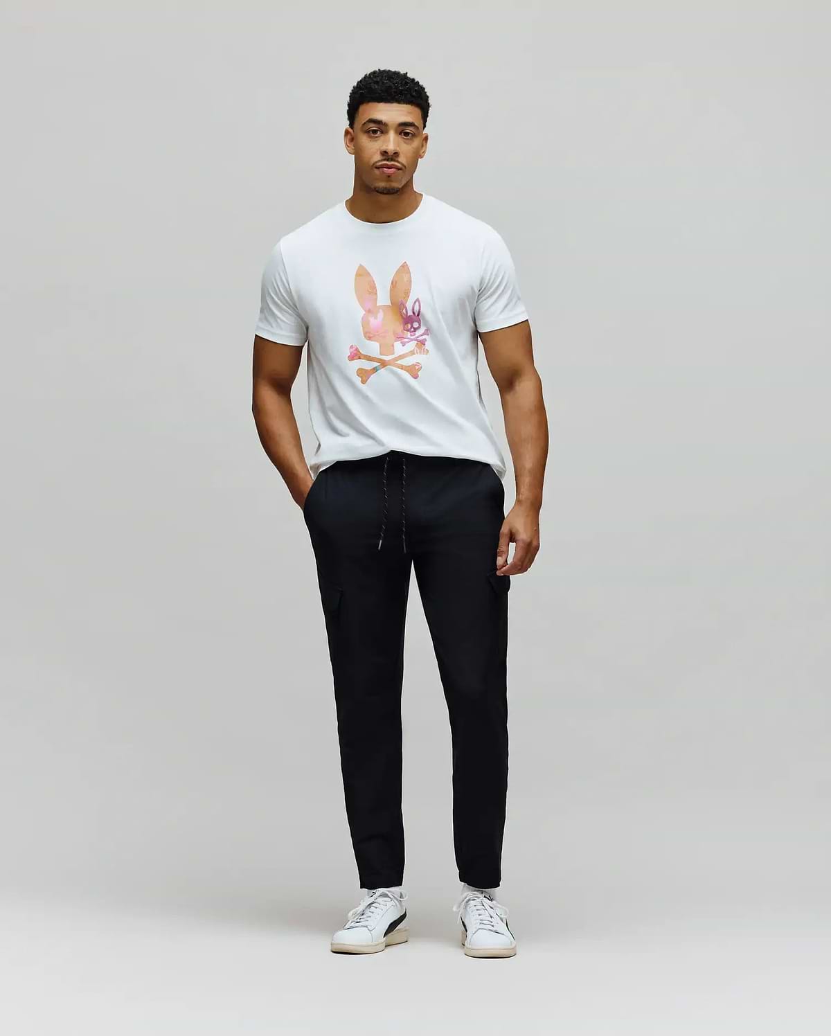 White classic fit jersey t-shirt with a large, colorful watercolor Psycho Bunny skull logo on the chest, paired with black slim fit Mens Luka Cargo Pant featuring a drawstring waist and side pockets.