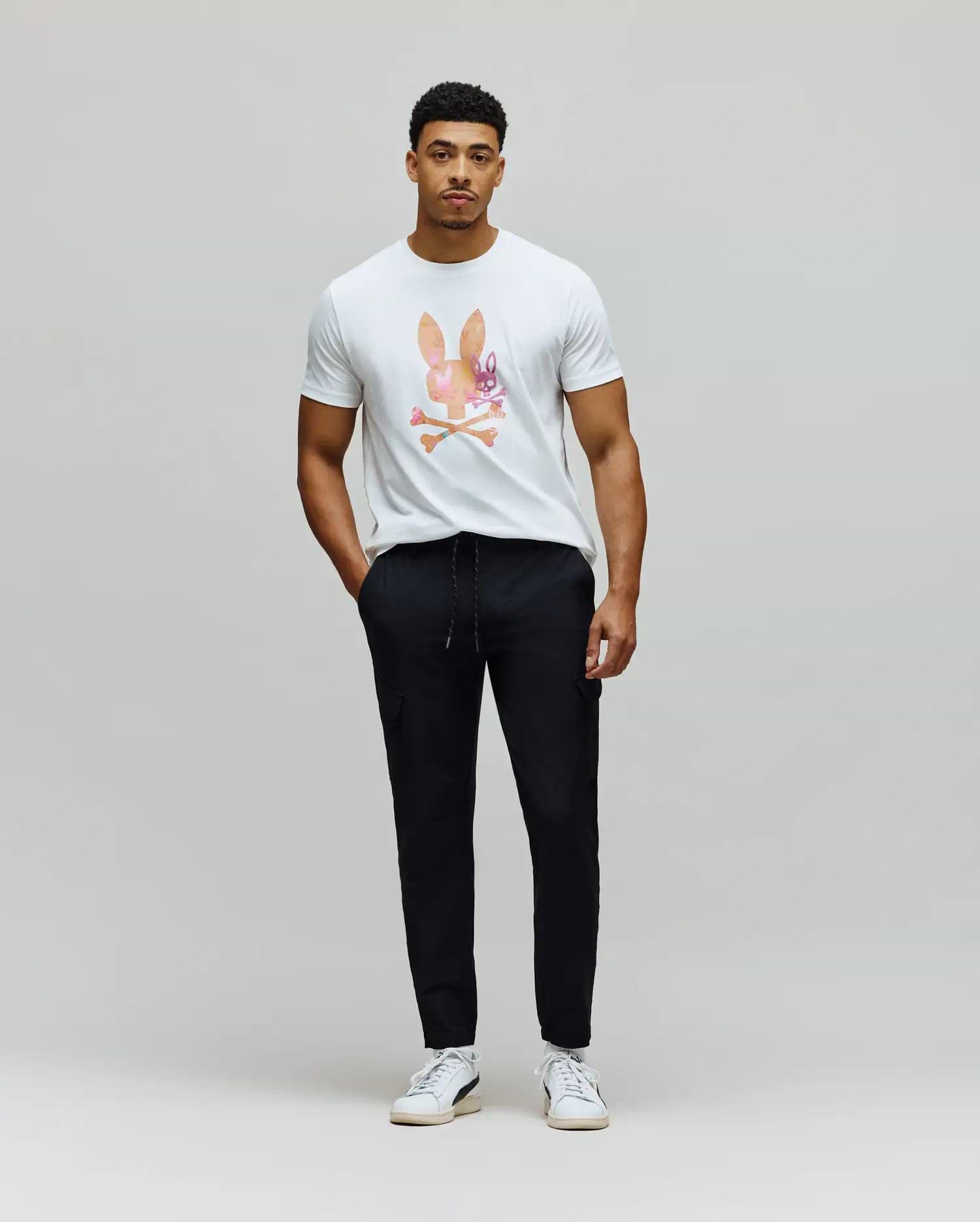 White classic fit jersey t-shirt with a large, colorful watercolor Psycho Bunny skull logo on the chest, paired with black slim fit Mens Luka Cargo Pant featuring a drawstring waist and side pockets.