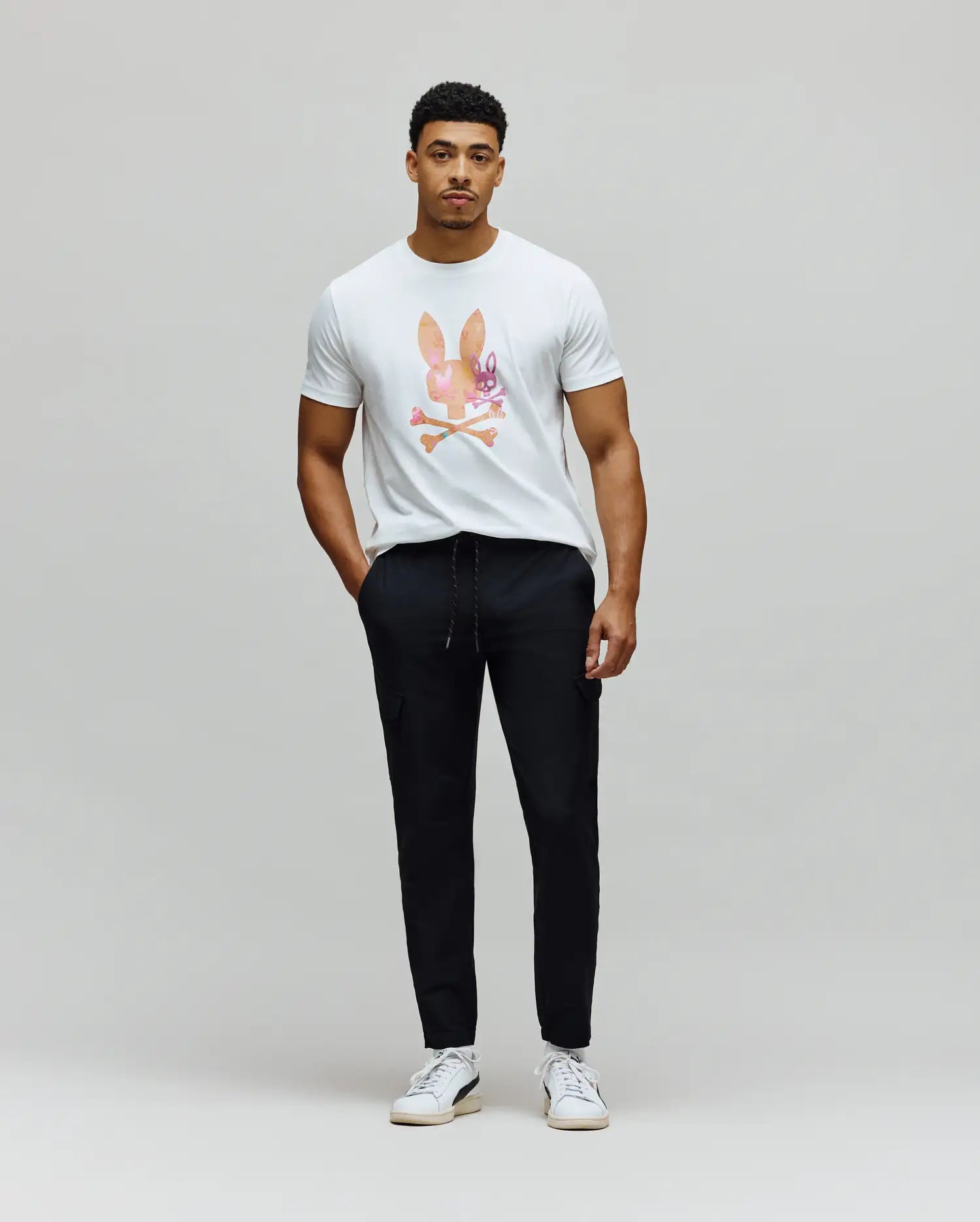 White classic fit jersey t-shirt with a large, colorful watercolor Psycho Bunny skull logo on the chest, paired with black slim fit Mens Luka Cargo Pant featuring a drawstring waist and side pockets.