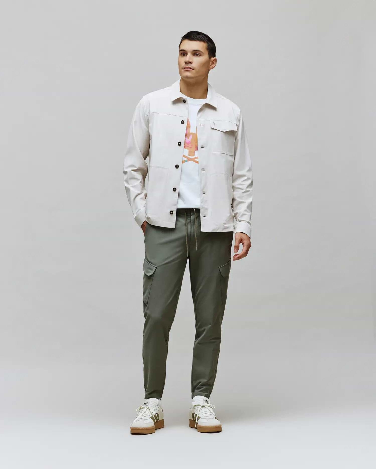 Mens Luka Cargo Pant in olive green, a white jersey t-shirt with a vibrant Psycho Bunny skull graphic, and a light beige classic fit overshirt with chest pockets.
