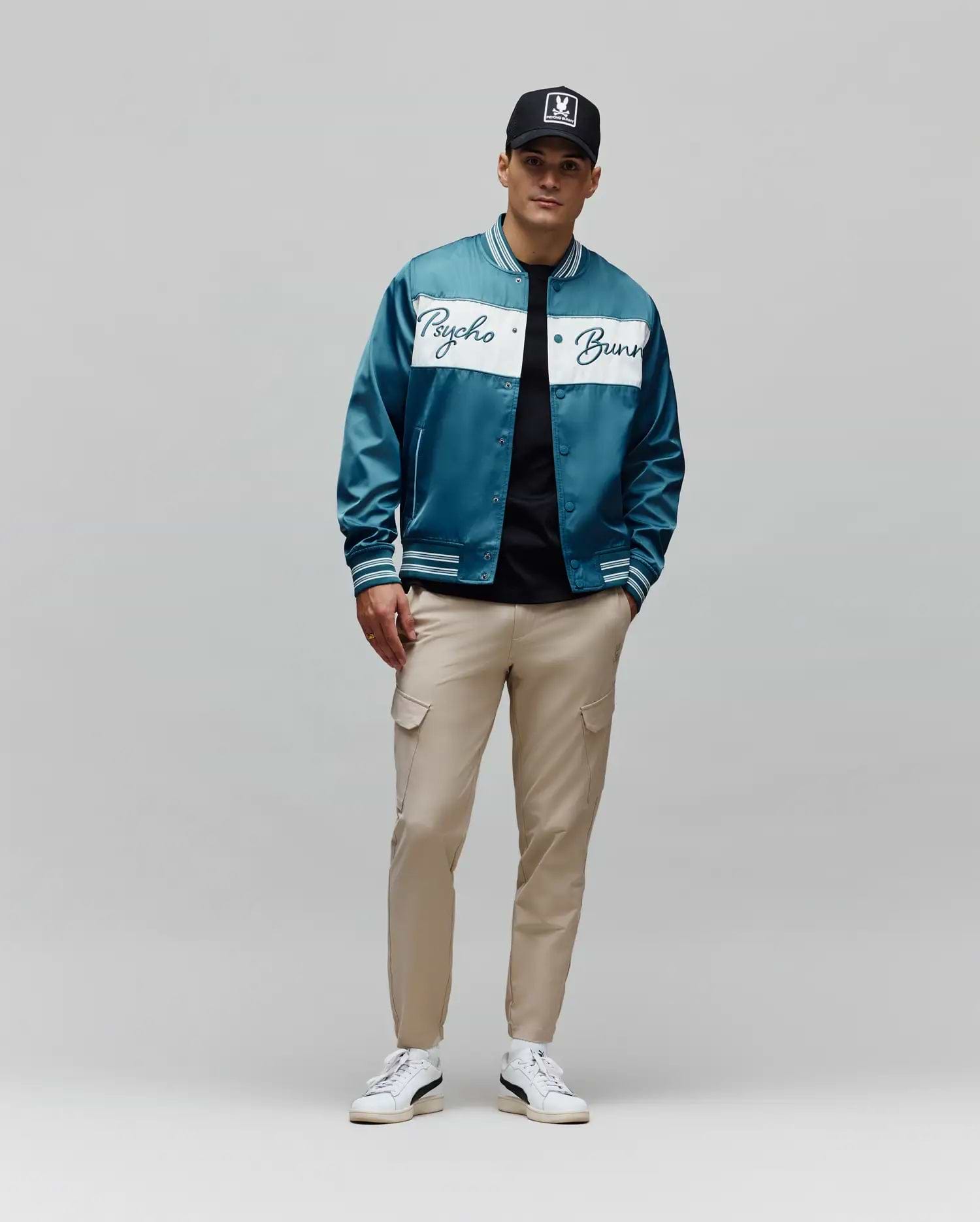 Psycho Bunny teal satin varsity jacket with white script logo and ribbed cuffs, classic fit black jersey tee, slim fit beige Mens Luka Cargo Pant, and black trucker cap with iconic white bunny skull logo.