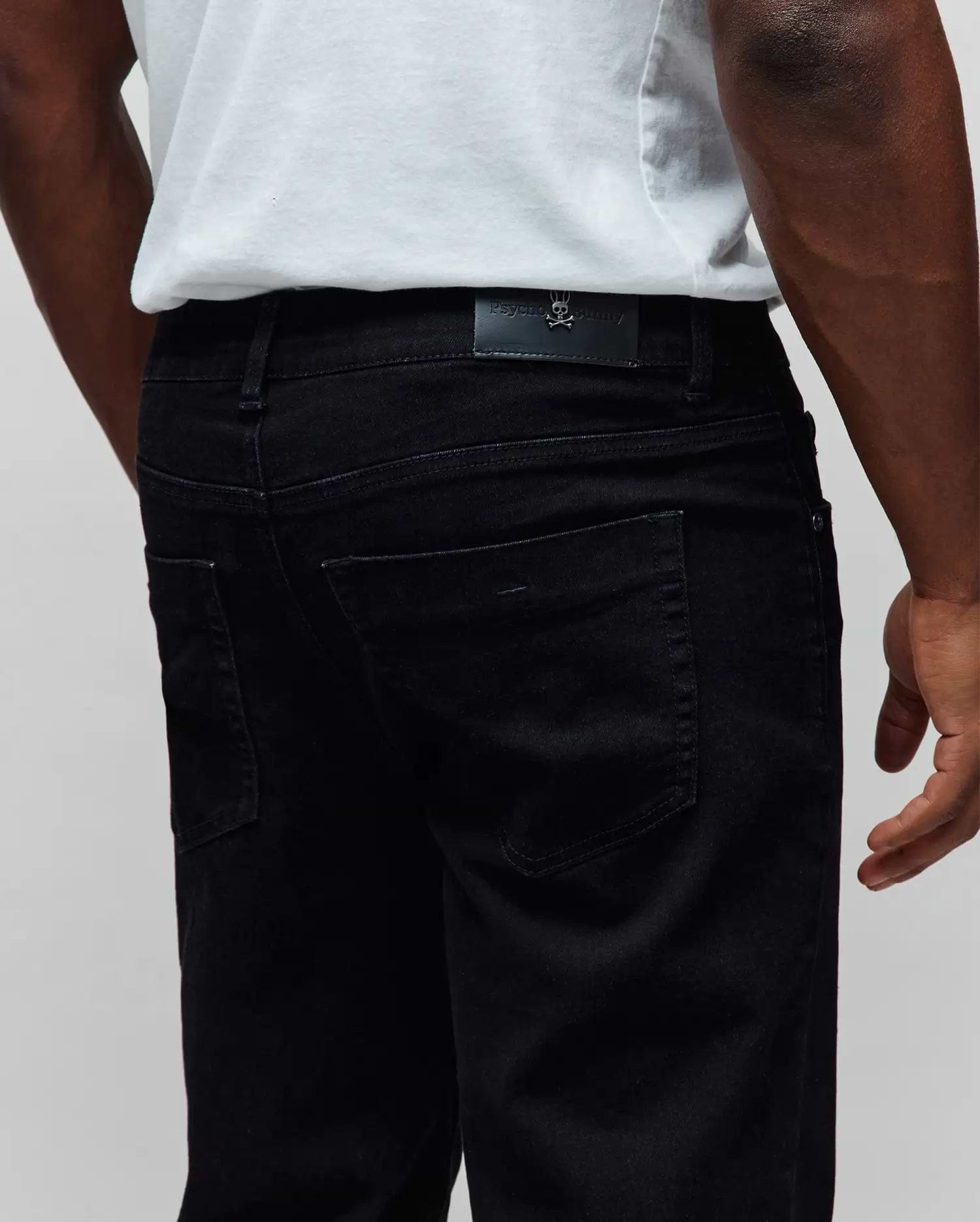 Black Mens Emerson Denim Pant, classic fit, with a black leather Psycho Bunny logo patch featuring a metallic skull on the back waistband, made from premium denim.