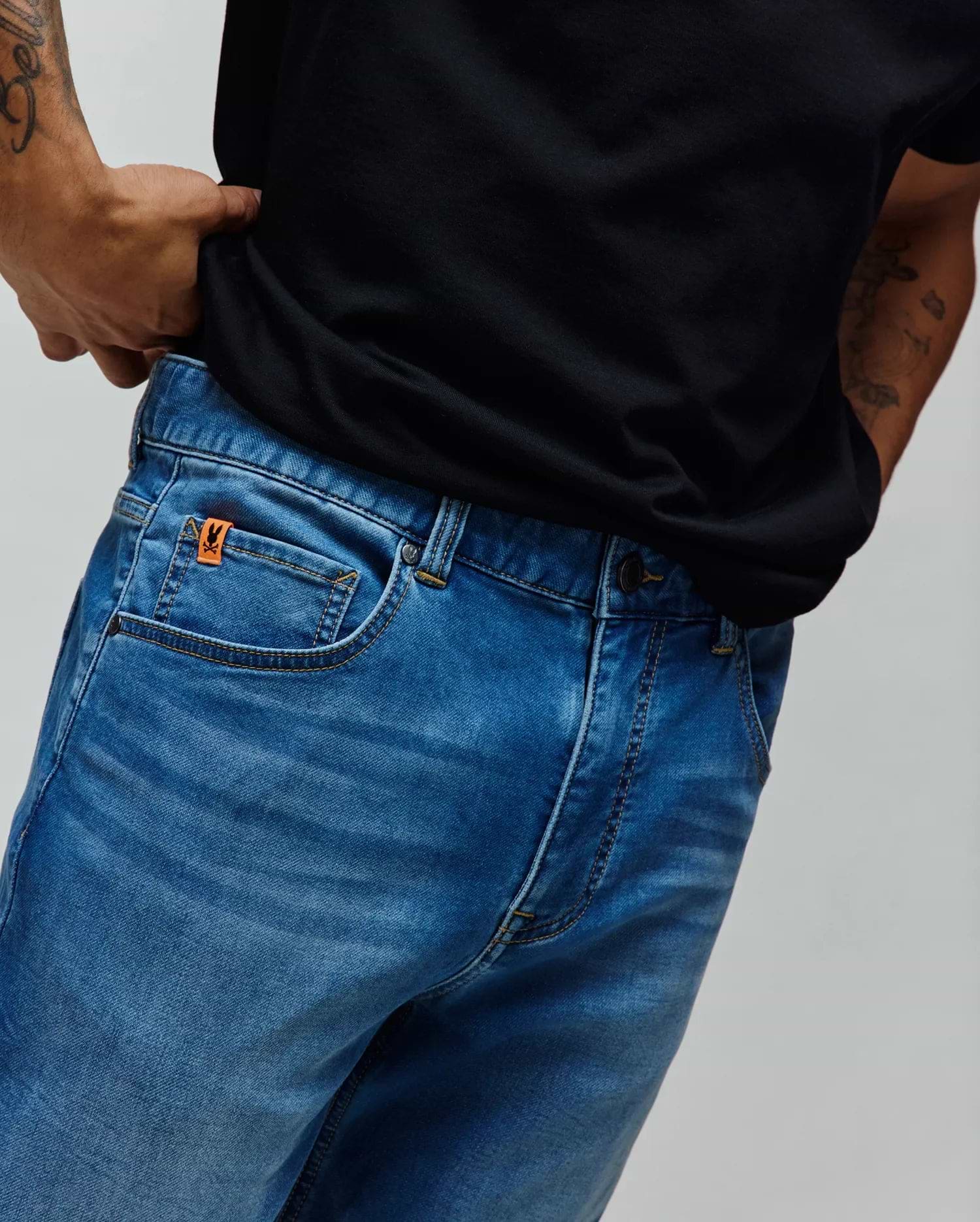 Mens Ace Knit Denim Pant in a classic blue wash with subtle fading, featuring contrast gold stitching and a small orange Psycho Bunny logo tag on the coin pocket.
