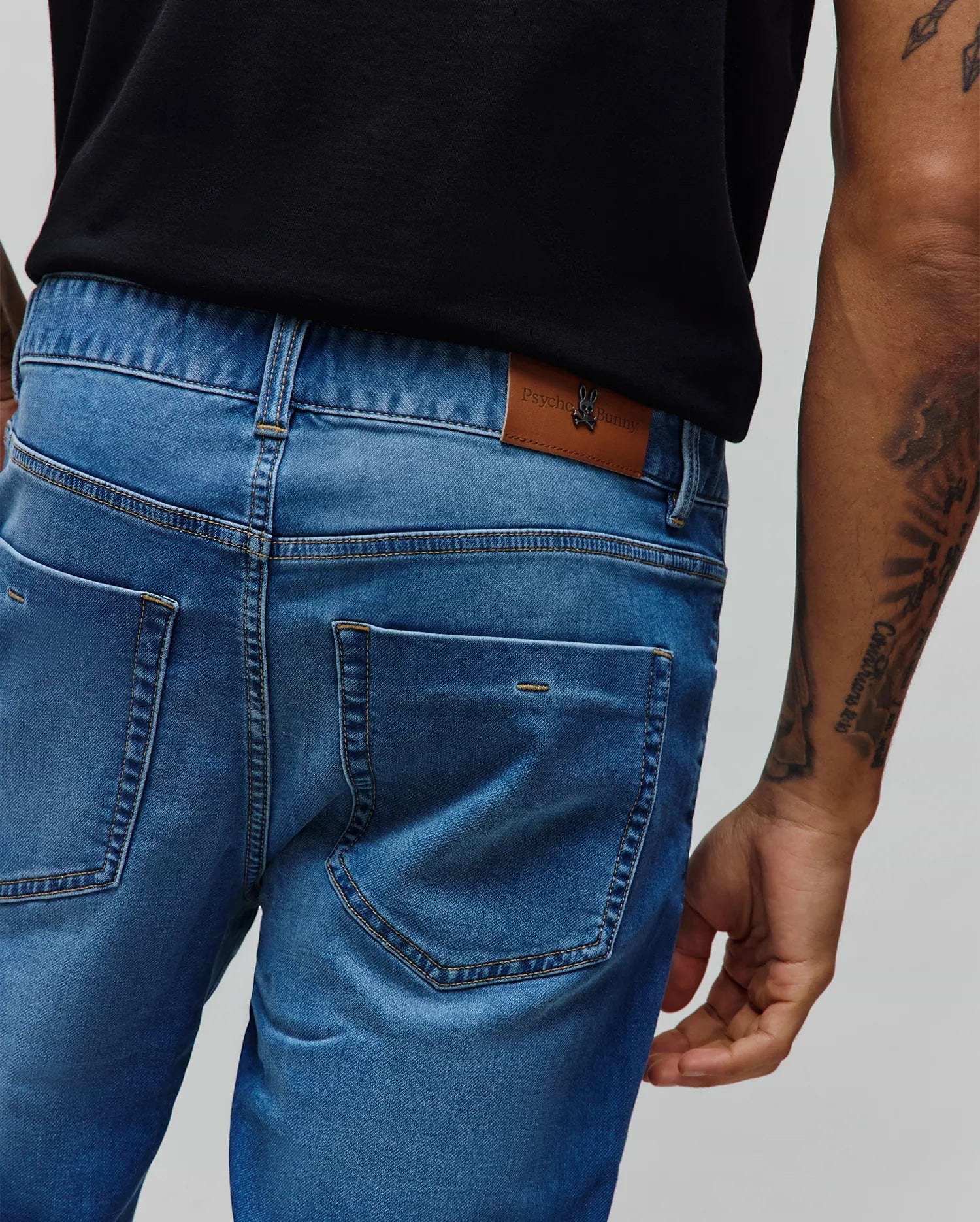 Mens Ace Knit Denim Pant in medium wash blue, featuring a classic fit, contrast stitching, and a tan Psycho Bunny logo patch with a metal bunny skull on the back waistband.