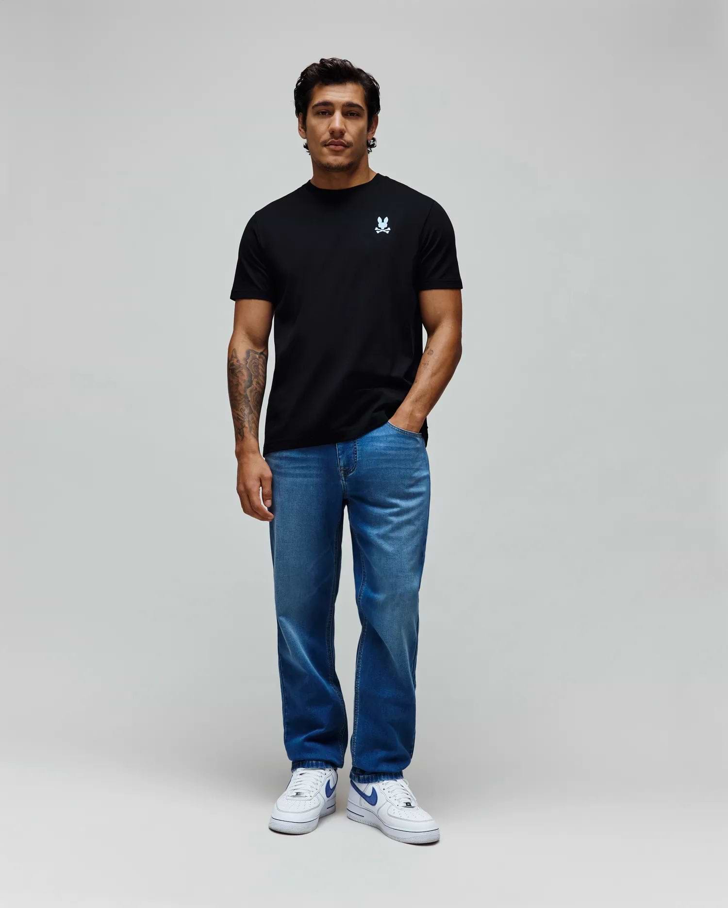 Black classic fit jersey t-shirt with a small embroidered light blue Psycho Bunny skull logo on the left chest, paired with relaxed fit medium blue washed MENS ACE KNIT DENIM PANT.