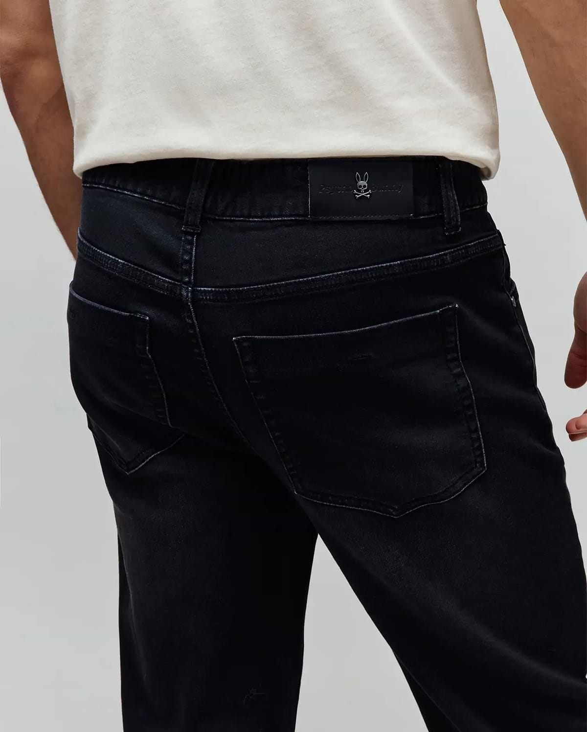 Mens Ace Knit Denim Pant. Washed black classic fit knit denim with back pockets, belt loops, and a small embossed Psycho Bunny skull logo on a black leather patch at the back waistband.