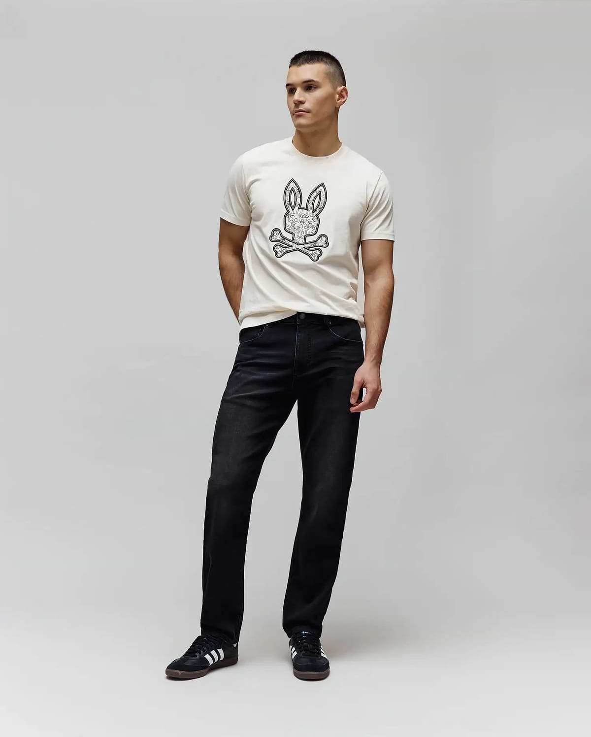 Cream jersey t-shirt with a large, intricate black Psycho Bunny skull logo on the chest, paired with dark washed black Mens Ace Knit Denim Pant in a relaxed fit.