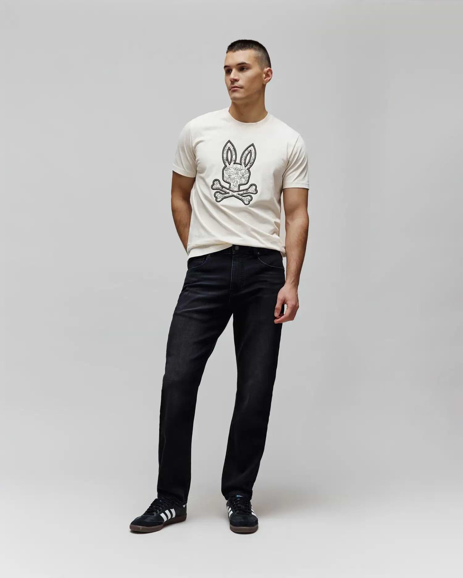 Cream jersey t-shirt with a large, intricate black Psycho Bunny skull logo on the chest, paired with dark washed black Mens Ace Knit Denim Pant in a relaxed fit.