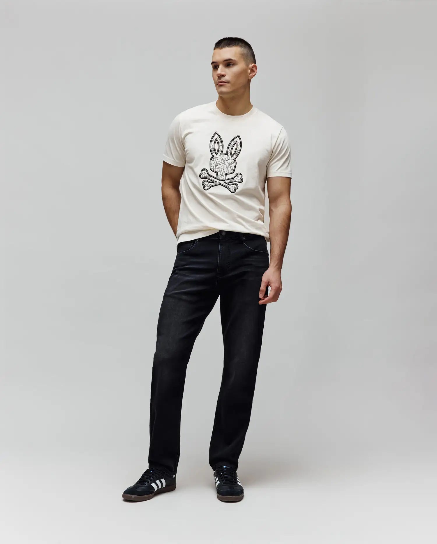 Cream jersey t-shirt with a large, intricate black Psycho Bunny skull logo on the chest, paired with dark washed black Mens Ace Knit Denim Pant in a relaxed fit.