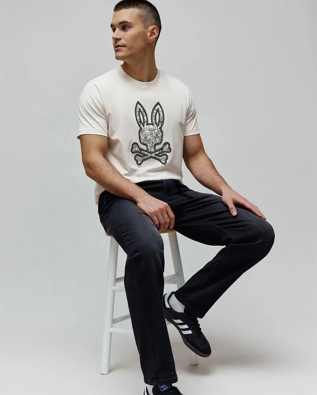 Cream classic fit jersey t-shirt with a large black and white intricate Psycho Bunny skull logo graphic on the chest, paired with dark grey Mens Ace Knit Denim Pant.