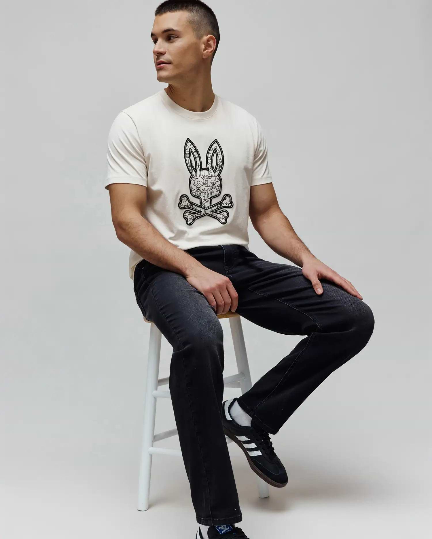 Cream classic fit jersey t-shirt with a large black and white intricate Psycho Bunny skull logo graphic on the chest, paired with dark grey Mens Ace Knit Denim Pant.
