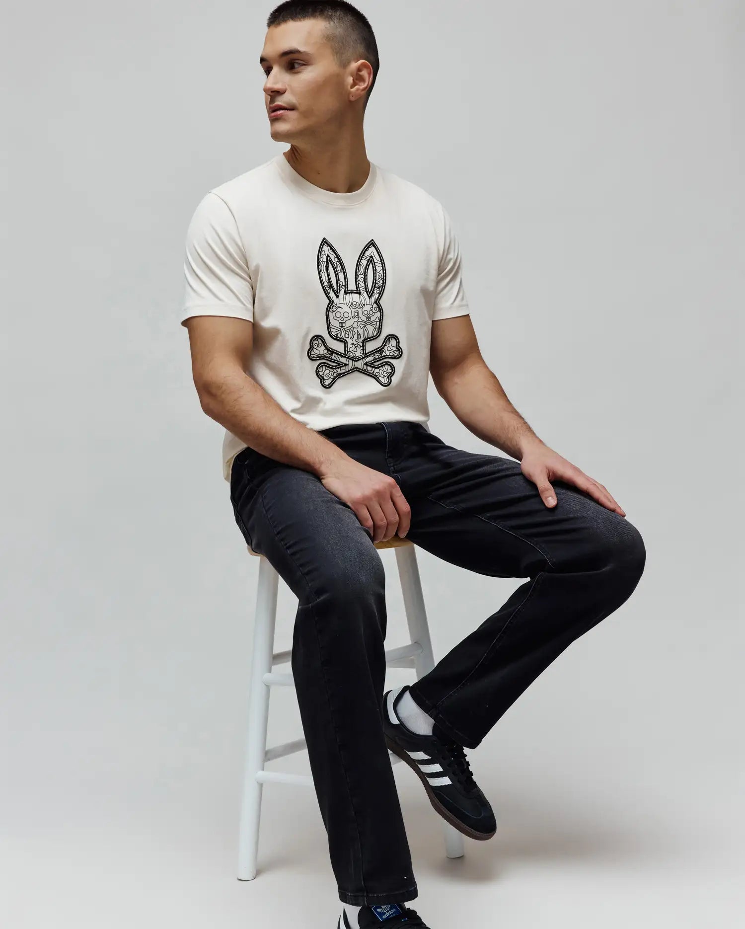 Cream classic fit jersey t-shirt with a large black and white intricate Psycho Bunny skull logo graphic on the chest, paired with dark grey Mens Ace Knit Denim Pant.
