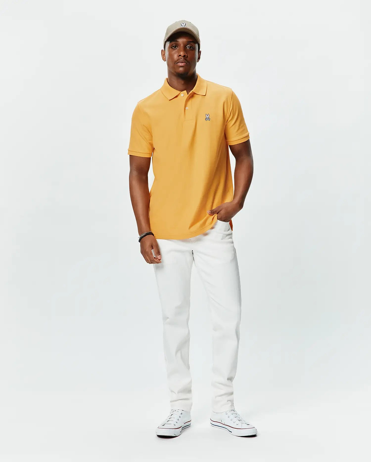 Orange pique polo shirt with classic fit and embroidered Psycho Bunny skull logo on left chest, paired with white slim-fit Mens Emerson Denim Pant and a beige cap with matching logo.