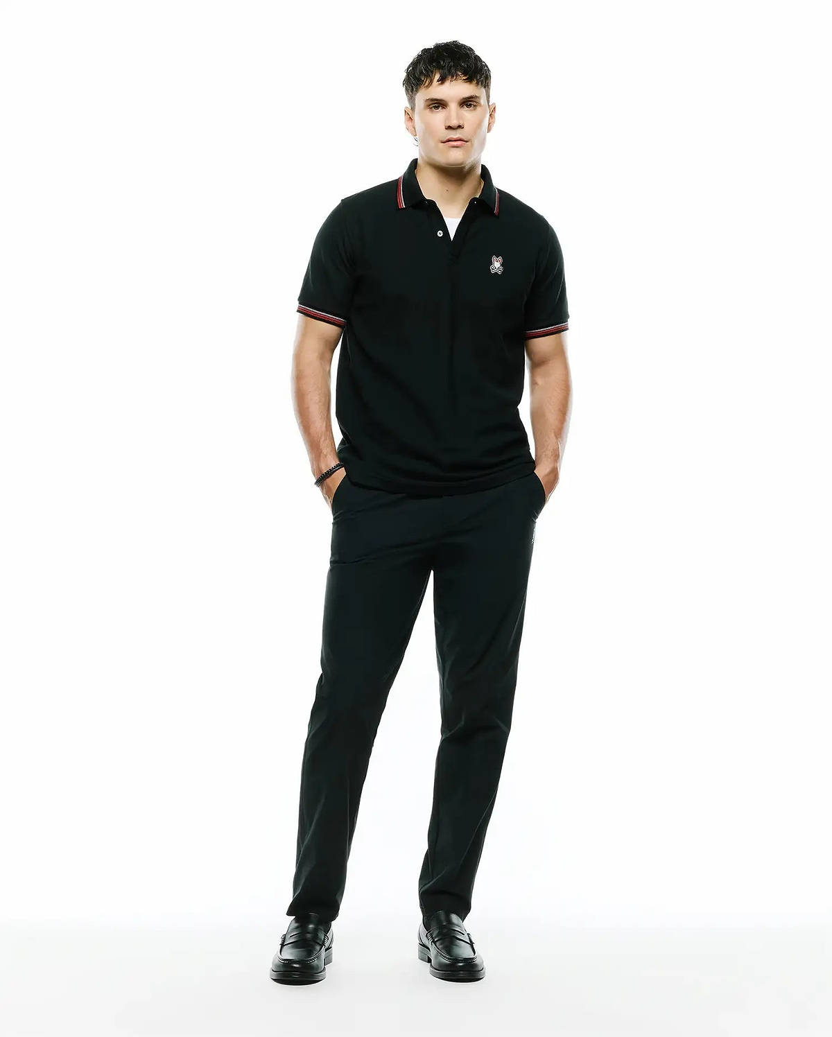 Black pique polo with red and white striped collar and cuffs, featuring an embroidered Psycho Bunny skull logo on the left chest, paired with black Mens Edward 10k Tapered Pant.