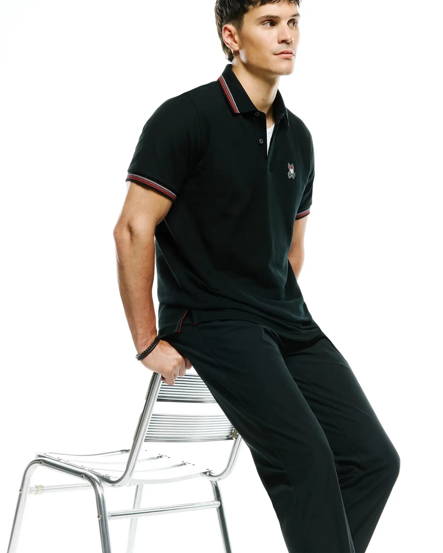 Black pique polo shirt with classic fit, featuring red and white striped collar and cuffs, and a small embroidered Psycho Bunny skull logo on the left chest. Paired with black Mens Edward 10k Tapered Pant.