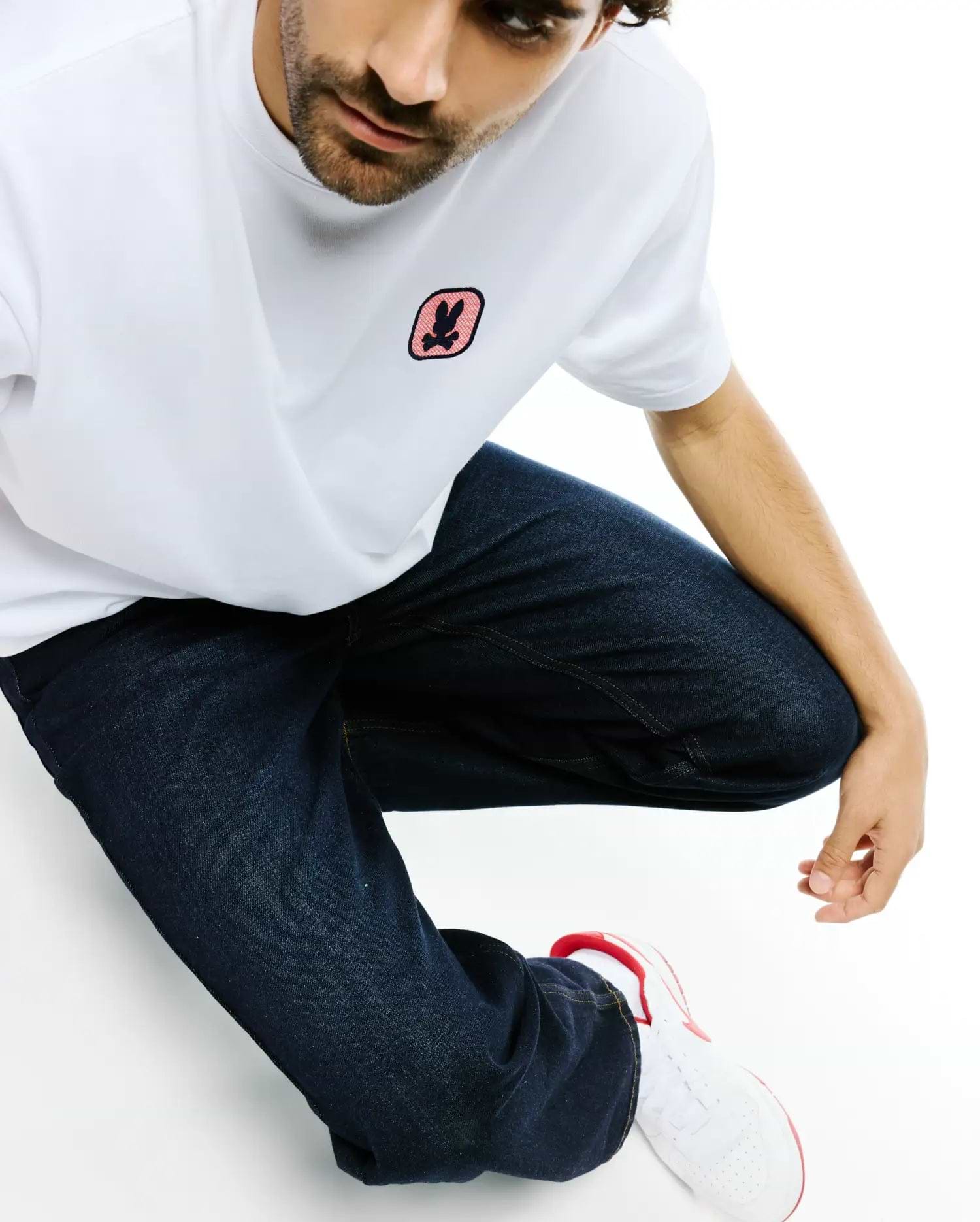 White classic fit jersey t-shirt with a small black and pink embroidered Psycho Bunny skull logo on the left chest, paired with dark blue relaxed fit denim Men's Beck Oversized Denim Pants. (3)