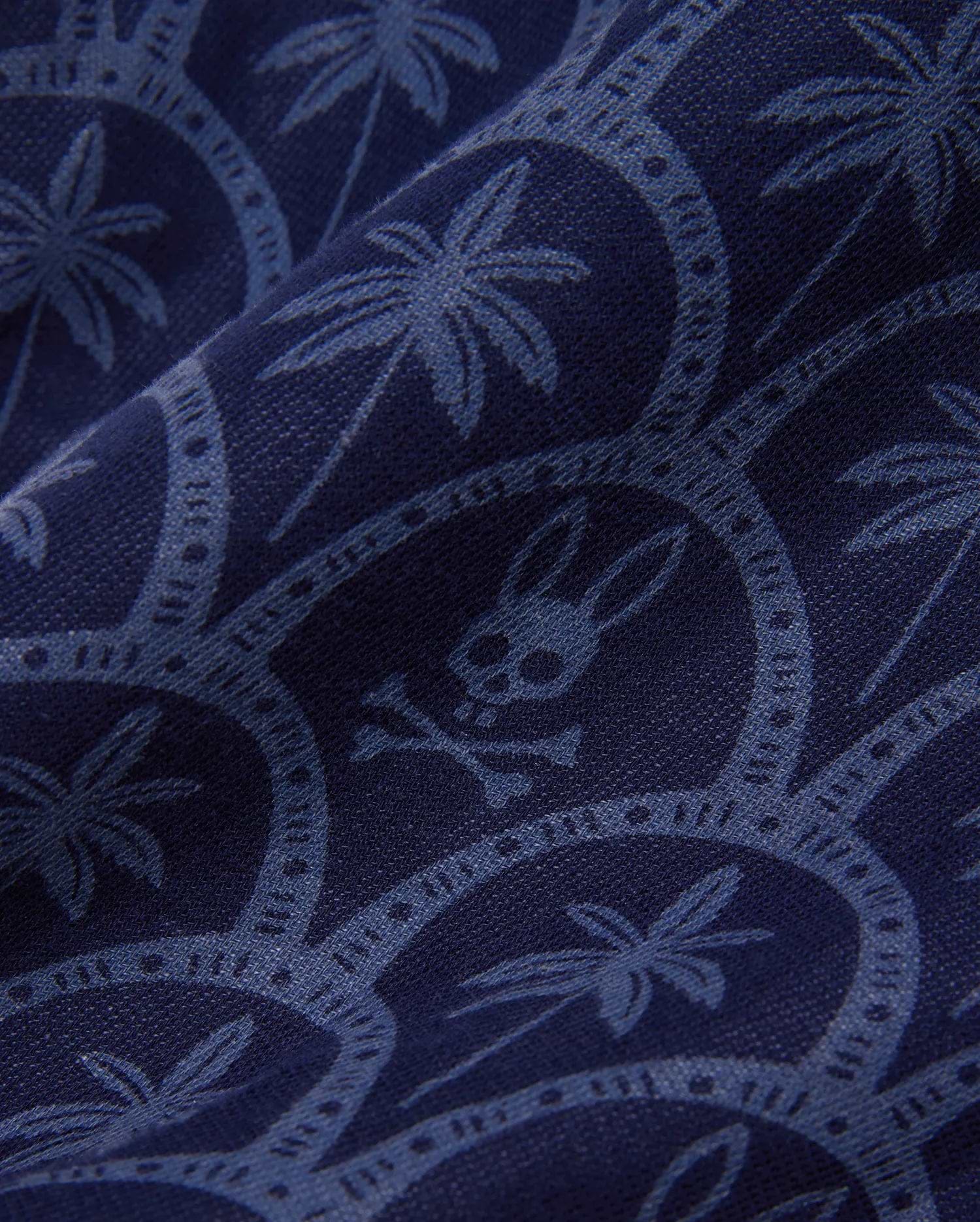 Mens Brad Linen Short 8 1/2 in navy blue, featuring a subtle lighter blue repeating print of palm trees, circular motifs, and the iconic Psycho Bunny skull logo on a woven linen fabric.