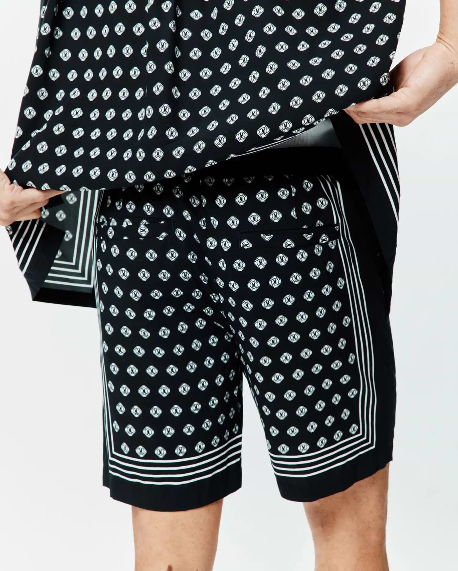 Mens Nick Tencel Short 7 in black, featuring a relaxed fit and smooth Tencel fabric. It has an all-over white Psycho Bunny skull logo diamond print, accented with bold white triple stripes along the side seams and hem.
