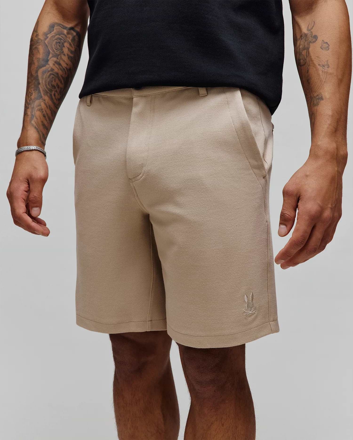 Mens Shiro Twill Short in classic fit beige twill fabric, featuring a subtle diagonal weave and a small, embroidered Psycho Bunny skull logo on the lower left leg.