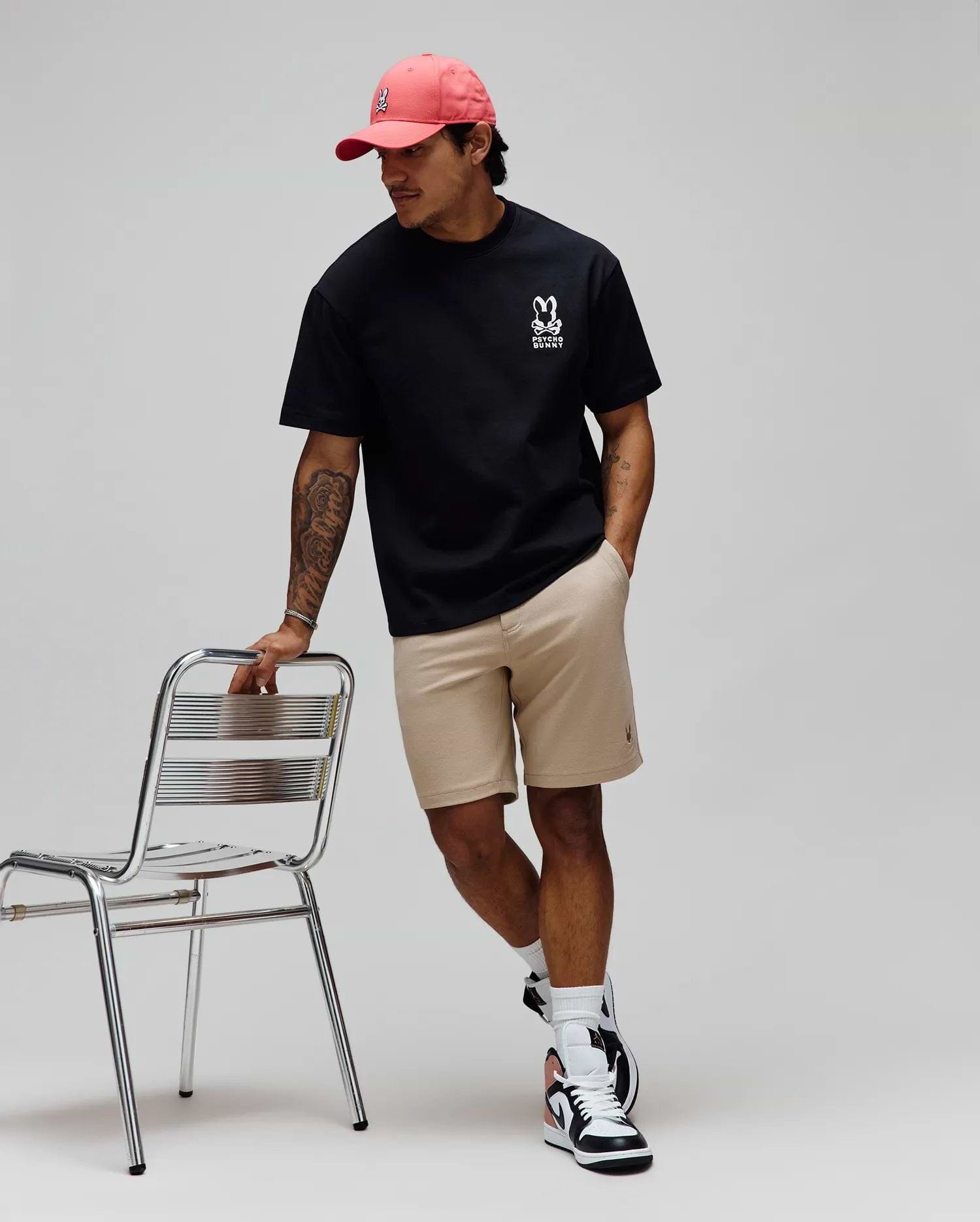 Pink cap with white embroidered Psycho Bunny skull, black relaxed-fit jersey tee with large white Psycho Bunny chest logo, and Khaki Mens Shiro Twill Short with subtle leg logo.