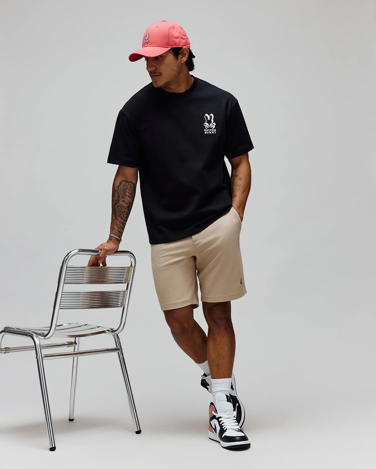 Pink cap with white embroidered Psycho Bunny skull, black relaxed-fit jersey tee with large white Psycho Bunny chest logo, and Khaki Mens Shiro Twill Short with subtle leg logo.