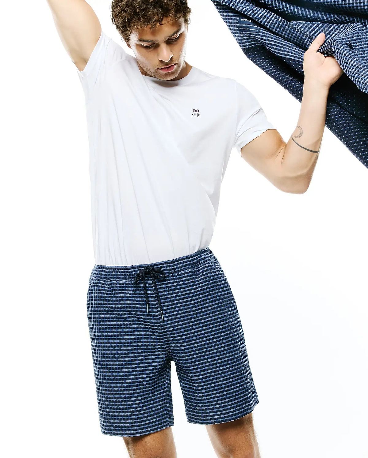 White jersey t-shirt with small embroidered Psycho Bunny skull logo on left chest, paired with Mens Erik Raised Check Short in navy and light blue, featuring a textured pattern and drawstring waist.