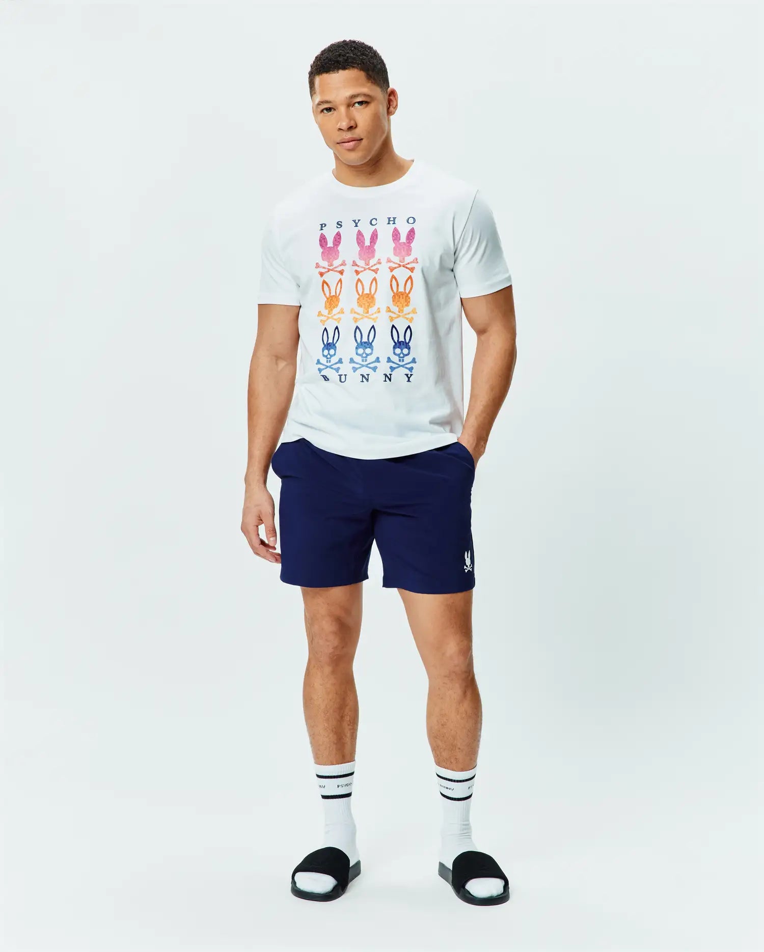 White jersey t-shirt with a large central graphic of nine gradient Psycho Bunny skull logos and PSYCHO BUNNY text, paired with navy Mens Blaze Sport Short with a small white embroidered Psycho Bunny logo on the left leg.