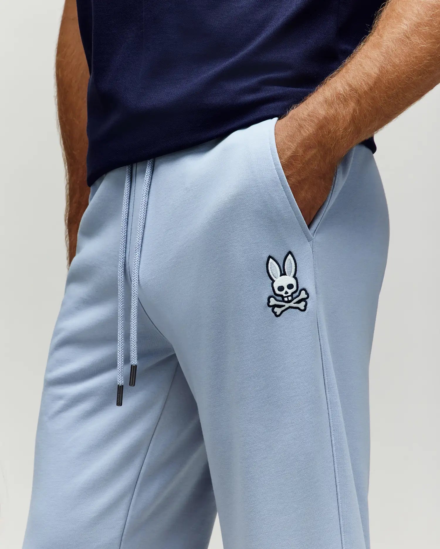 Mens Timothy 4k Bunny Jogger in light blue, featuring a relaxed fit, smooth French terry fabric, drawstring waist with dark tips, and an embroidered white and black Psycho Bunny skull logo on the left thigh.
