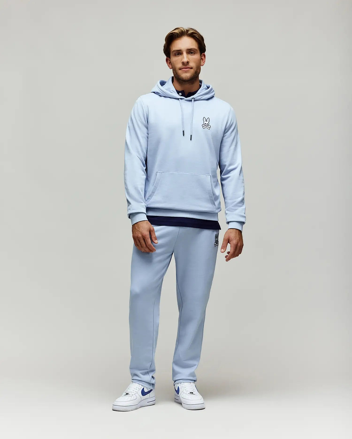 Light blue relaxed fit French terry hoodie with small embroidered Psycho Bunny skull logo on chest, kangaroo pocket, and matching Mens Timothy 4k Bunny Jogger with larger embroidered logo on thigh.