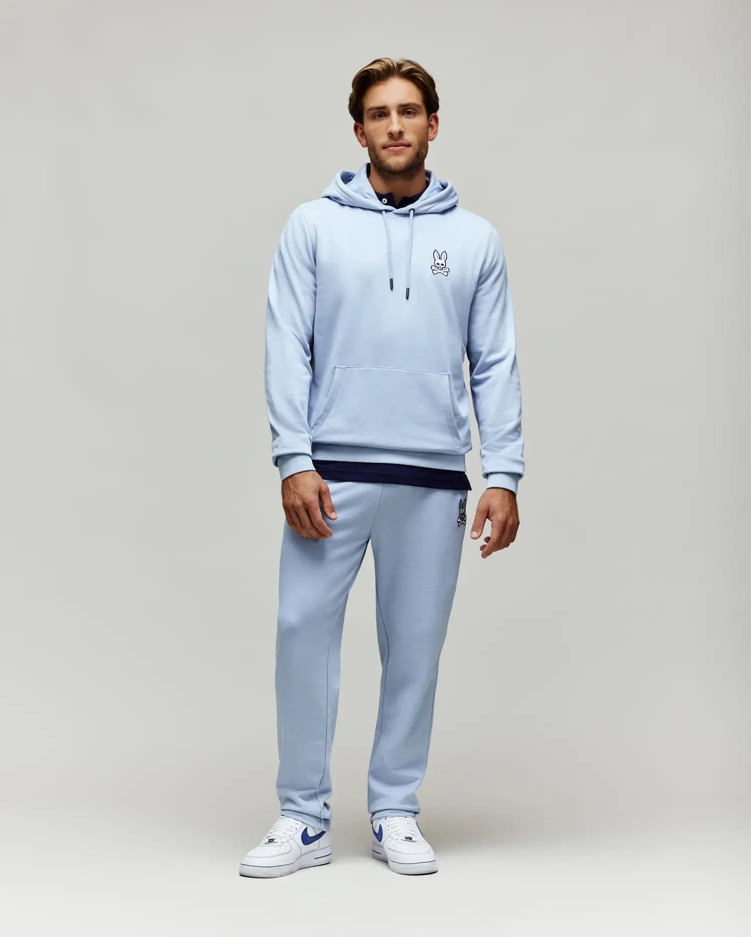 Light blue relaxed fit French terry hoodie with small embroidered Psycho Bunny skull logo on chest, kangaroo pocket, and matching Mens Timothy 4k Bunny Jogger with larger embroidered logo on thigh.