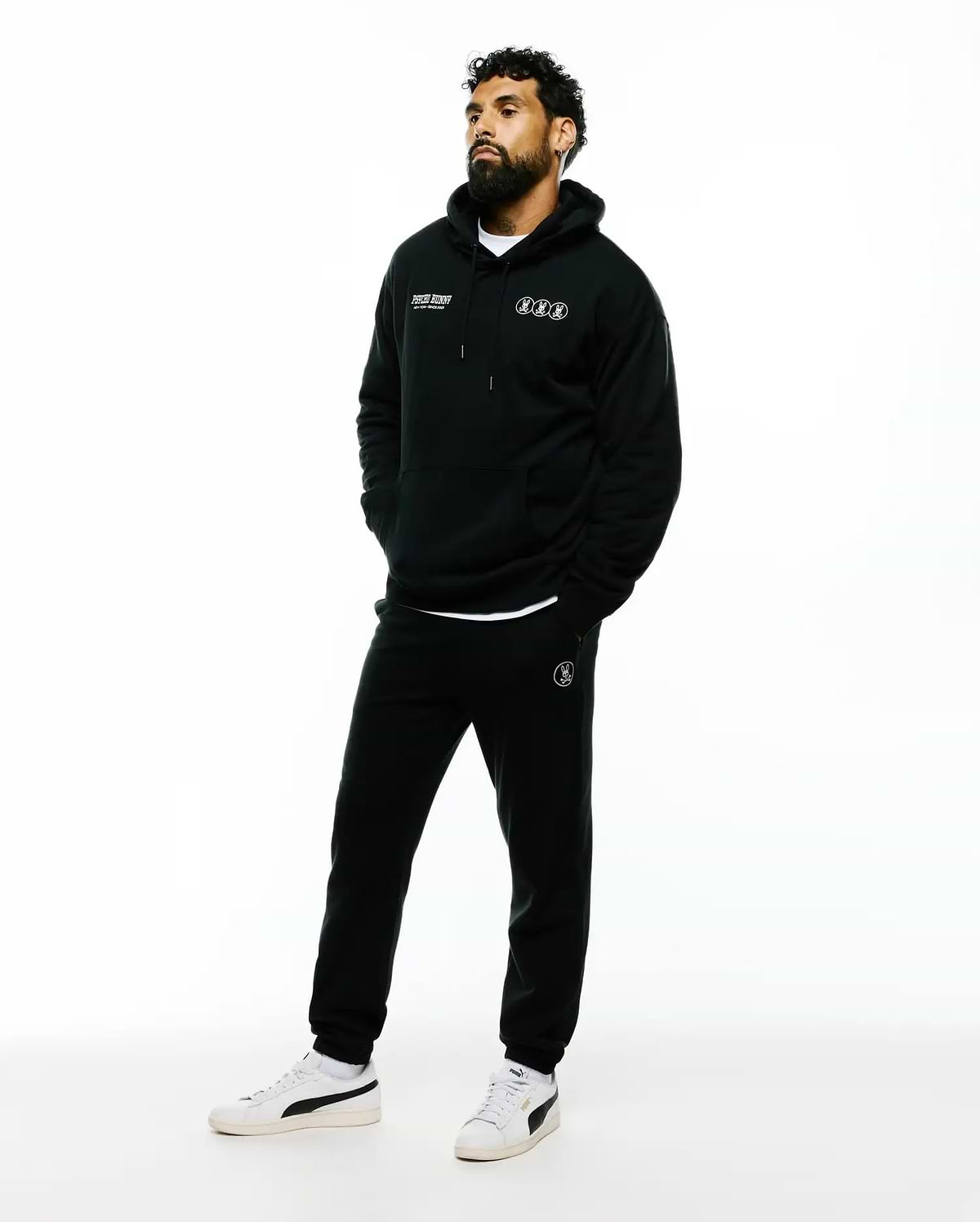 Black relaxed fit French terry hoodie with white Psycho Bunny text and three circular bunny skull logos on chest, paired with Mens Nathaniel Sweatpant featuring a white circular bunny skull logo on left thigh.