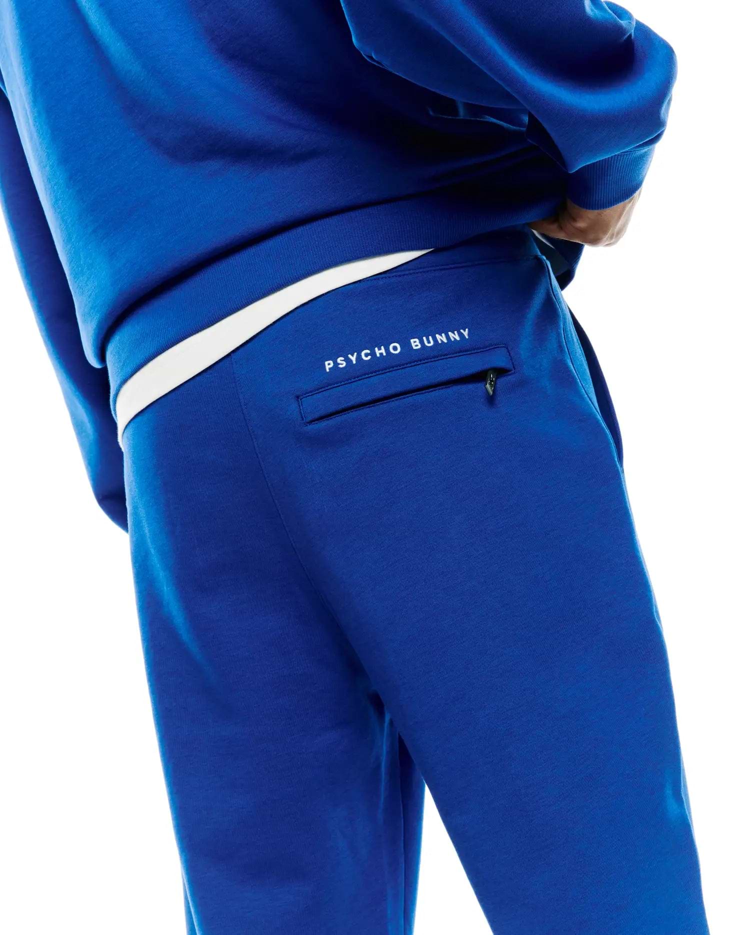 Royal blue Mens Nathaniel Sweatpant, a relaxed fit french terry garment, features a white Psycho Bunny text logo above a zippered back pocket and a contrasting white waistband.