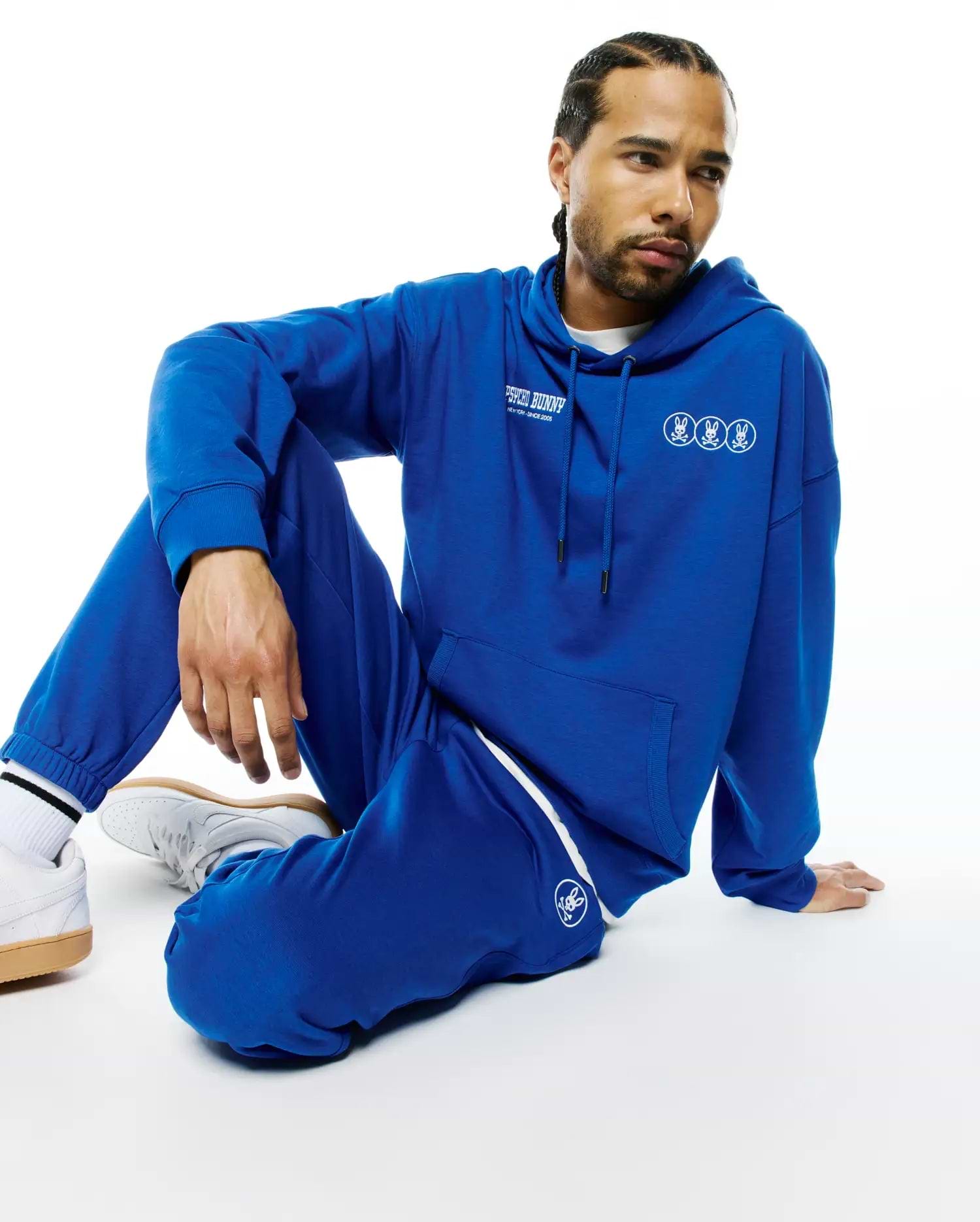 Royal blue relaxed fit French terry hoodie with white Psycho Bunny text and triple bunny skull chest logos, paired with matching Mens Nathaniel Sweatpant featuring a white side stripe and left thigh bunny skull logo.