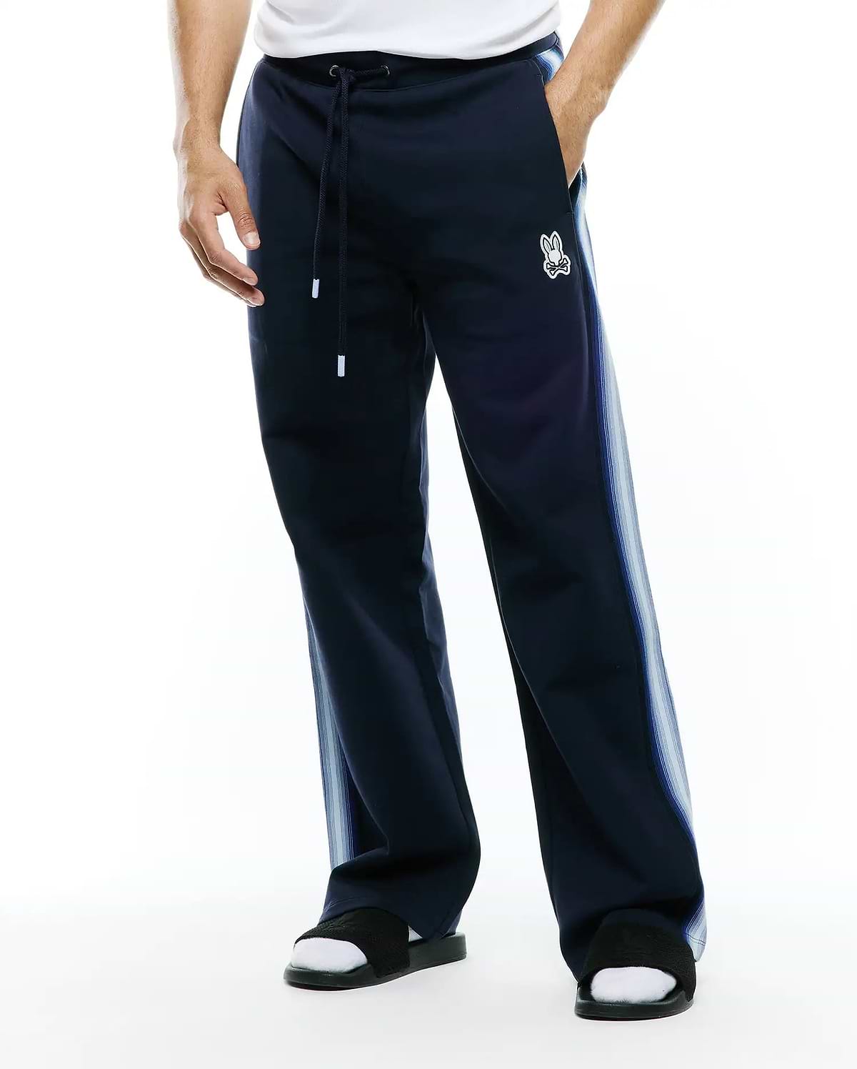 Mens Jaydon Track Pant in navy, featuring a relaxed fit, smooth knit fabric, drawstring waist, and a small embroidered white Psycho Bunny skull logo on the left thigh, with light blue gradient vertical stripes on the sides. (1)