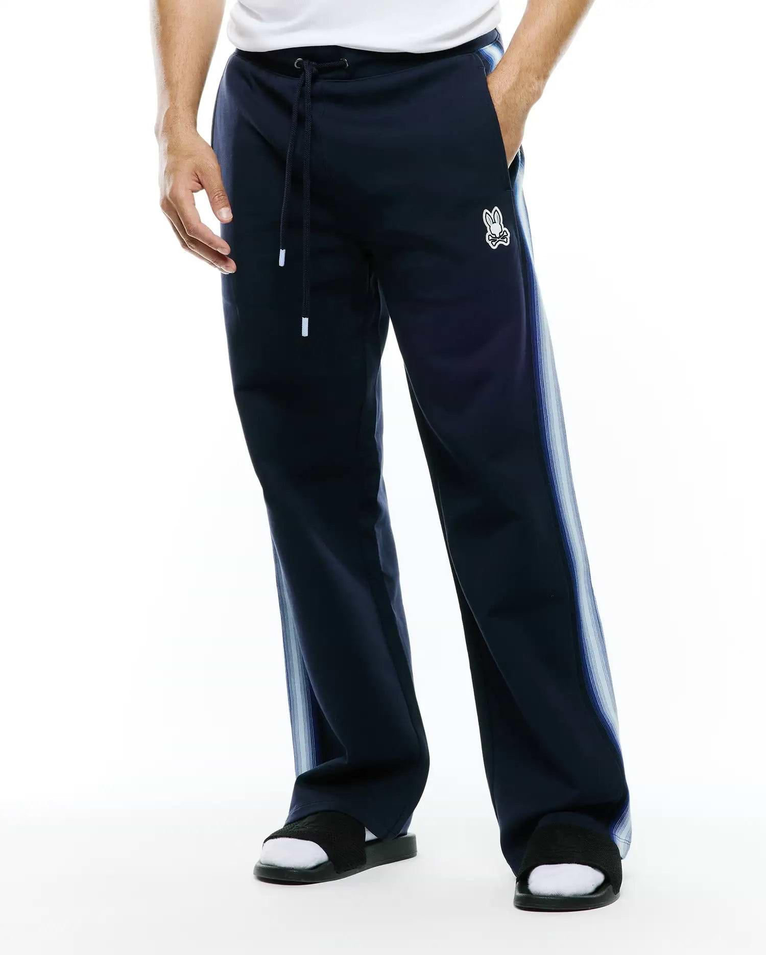 Mens Jaydon Track Pant in navy, featuring a relaxed fit, smooth knit fabric, drawstring waist, and a small embroidered white Psycho Bunny skull logo on the left thigh, with light blue gradient vertical stripes on the sides. (1)