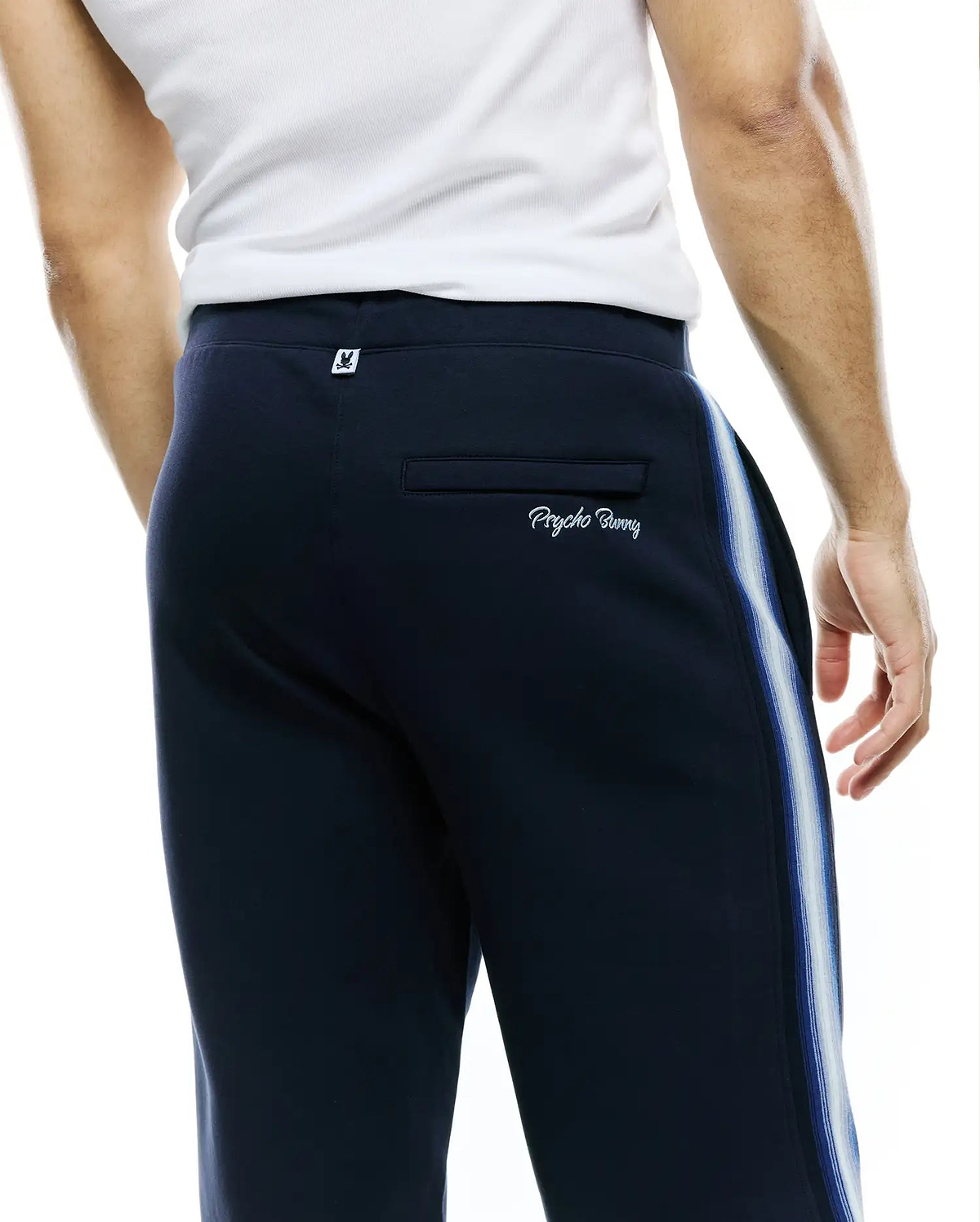 Mens Jaydon Track Pant, navy, relaxed fit jersey, featuring blue/white side stripes, small bunny skull logo tag on back waistband, and embroidered Psycho Bunny wordmark on back pocket. (3)