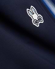 Navy blue Mens Jaydon Track Pant in smooth knit, featuring a prominent embroidered white and light blue Psycho Bunny skull logo and a multi-tonal blue vertical side stripe. (6)