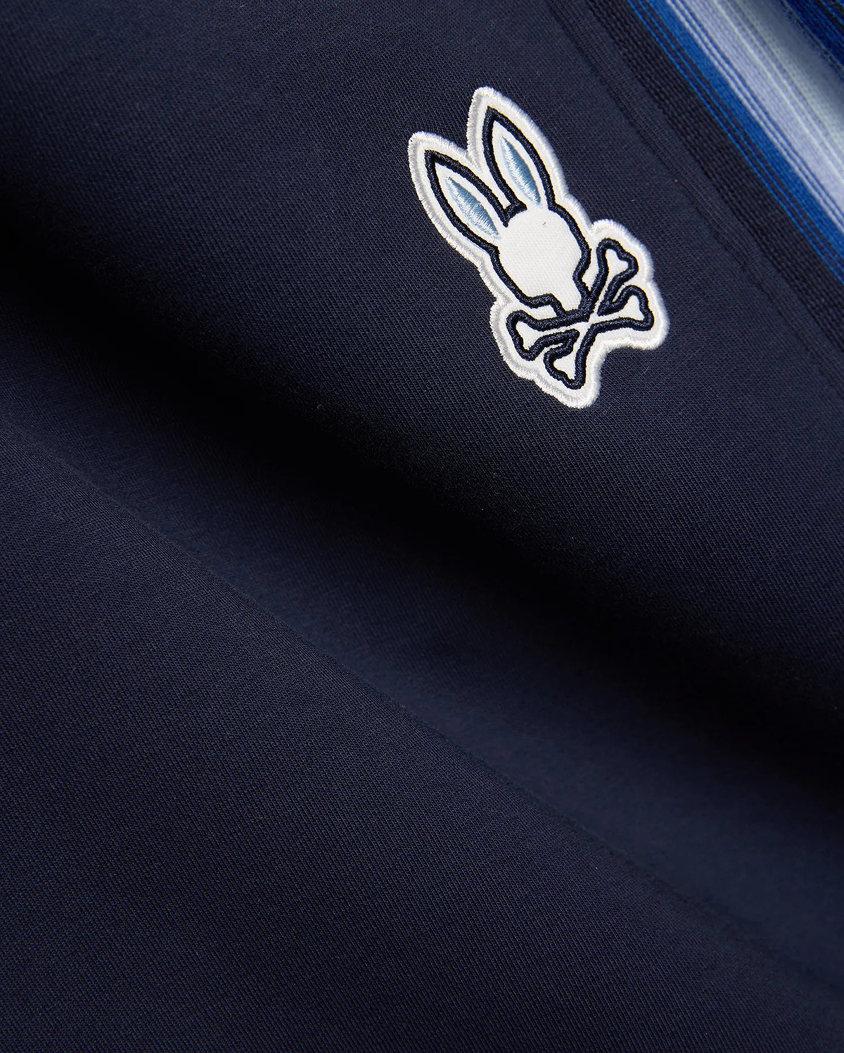 Navy blue Mens Jaydon Track Pant in smooth knit, featuring a prominent embroidered white and light blue Psycho Bunny skull logo and a multi-tonal blue vertical side stripe. (6)