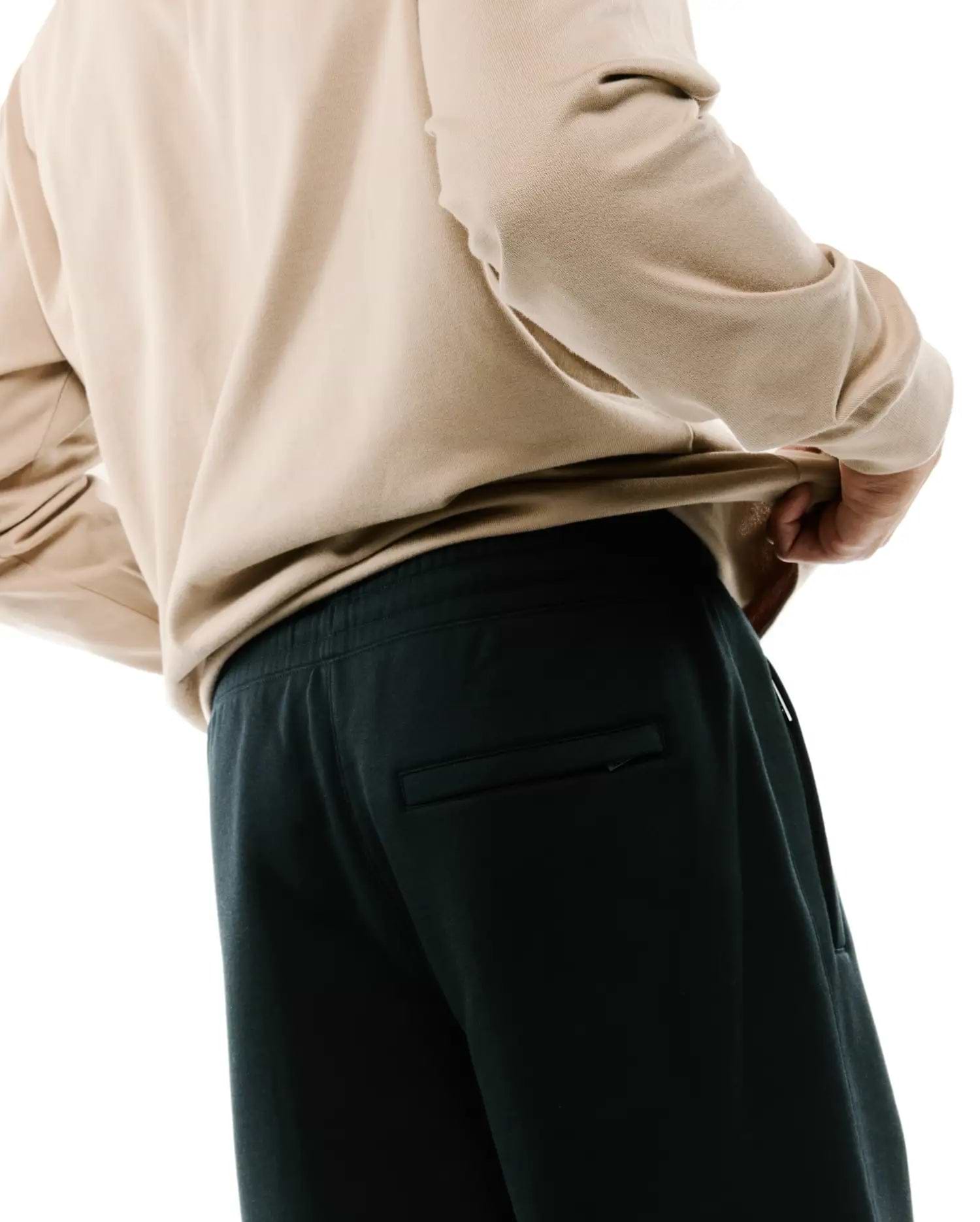 Dark charcoal Mens Terrence Brushed Fleece Sweatpant featuring a relaxed fit, elastic waistband, side pockets, and a subtle zippered back pocket, paired with a beige long-sleeve top.