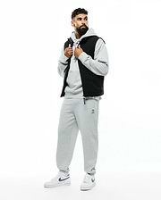 Mens Terrence Brushed Fleece Sweatpant in heather grey with a small embroidered Psycho Bunny skull logo and blue drawstring detail, paired with a matching grey relaxed-fit hoodie and a black puffer vest with navy lining.