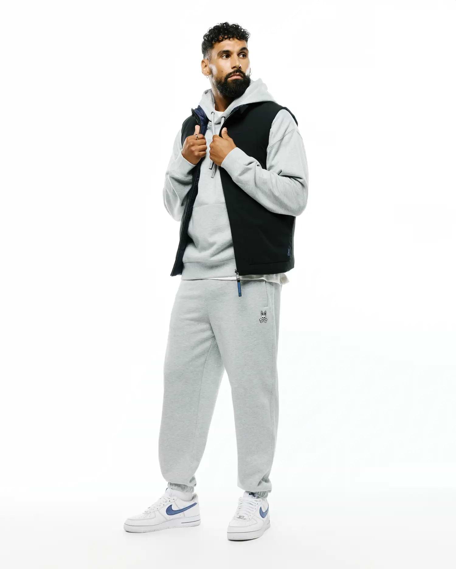 Mens Terrence Brushed Fleece Sweatpant in heather grey with a small embroidered Psycho Bunny skull logo and blue drawstring detail, paired with a matching grey relaxed-fit hoodie and a black puffer vest with navy lining.